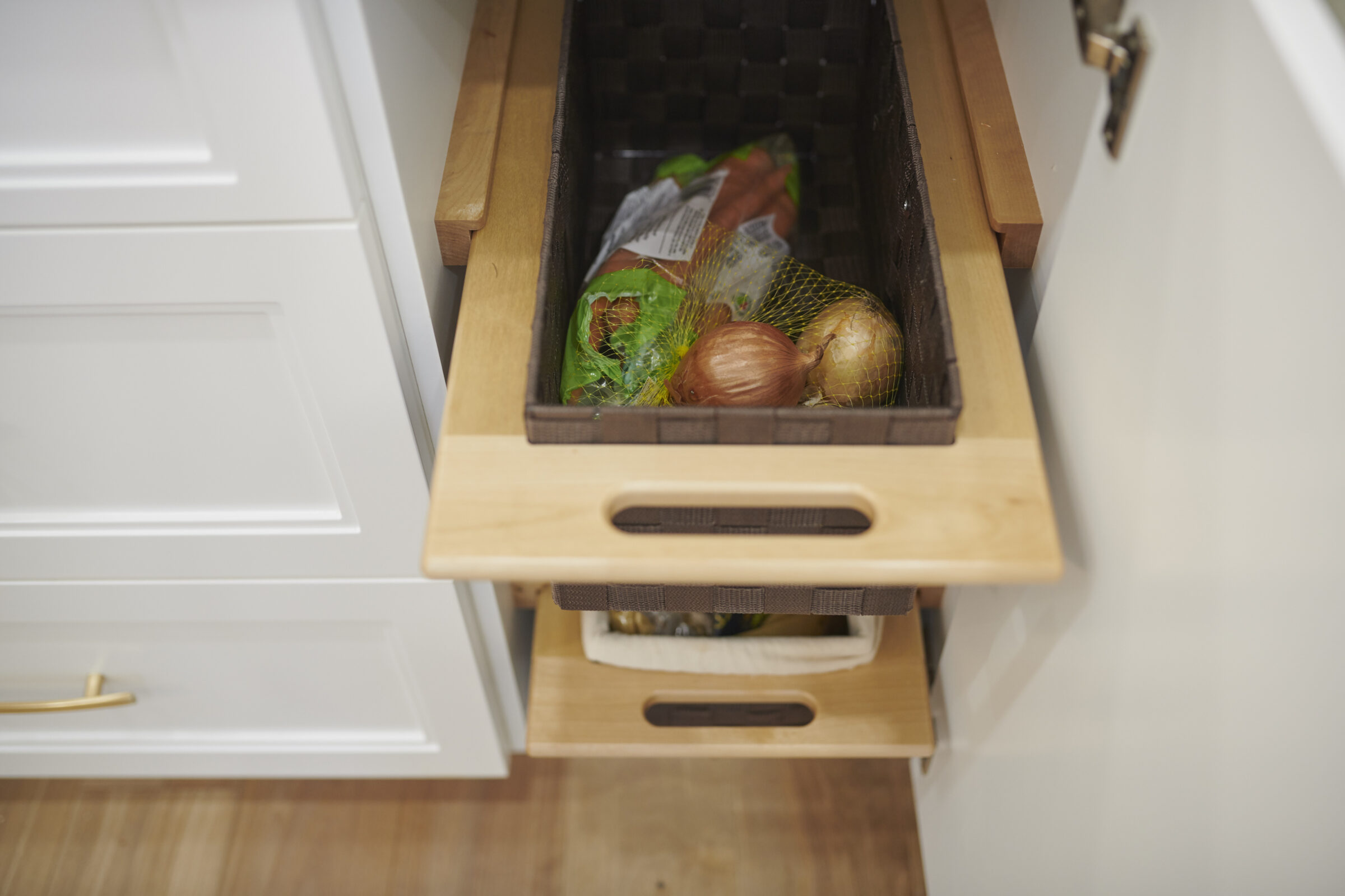 Two wooden kitchen drawers are partially open, revealing fresh produce like onions. The cabinet and handles have a modern design, with light-colored wood.