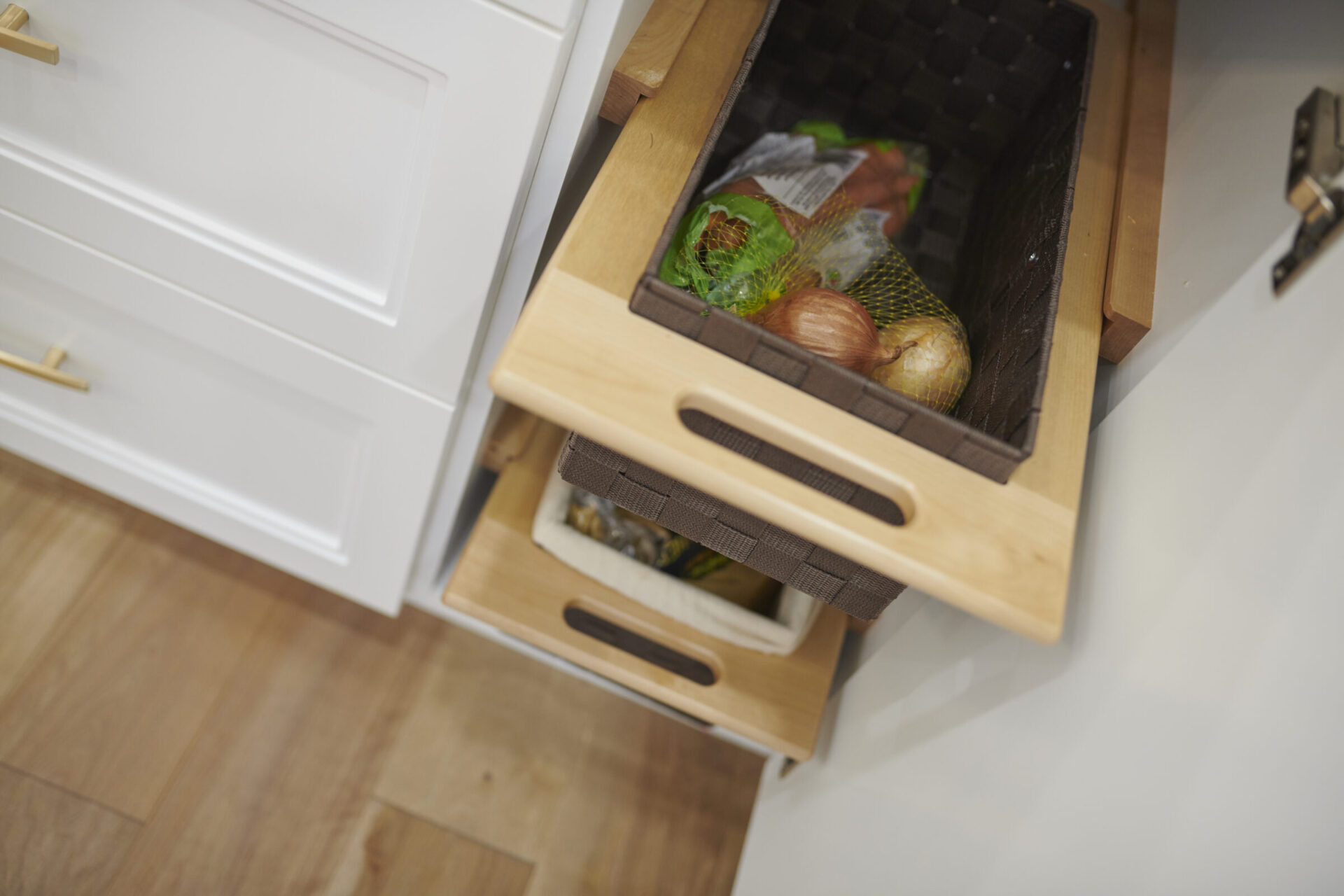 Two open wooden kitchen drawers with built-in baskets contain onions, potatoes, and a broccoli head. The modern design suggests an organized, clean space.