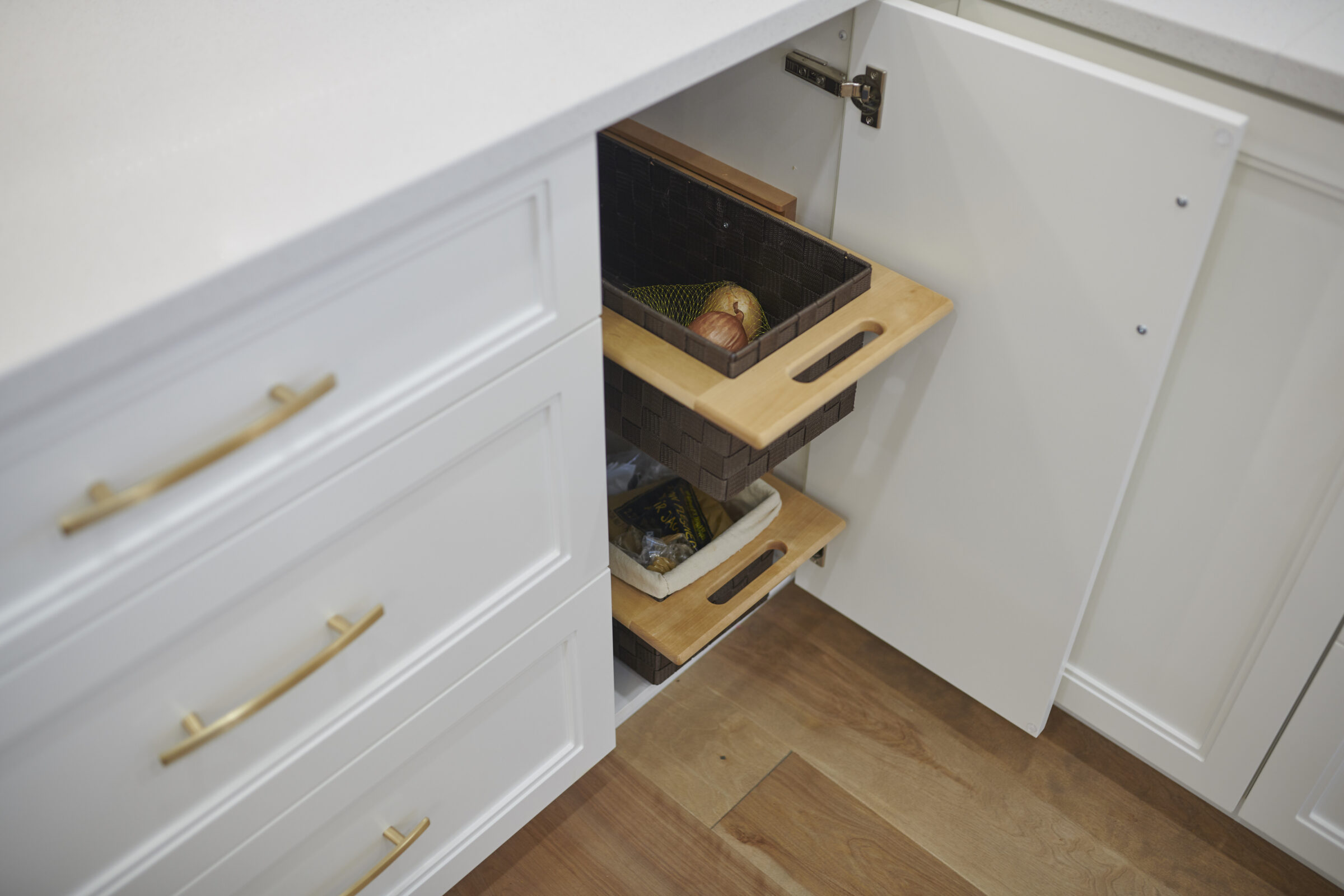 A kitchen cabinet with two pull-out wooden baskets is open, revealing stored food items, such as bread. The drawers have sleek gold handles.