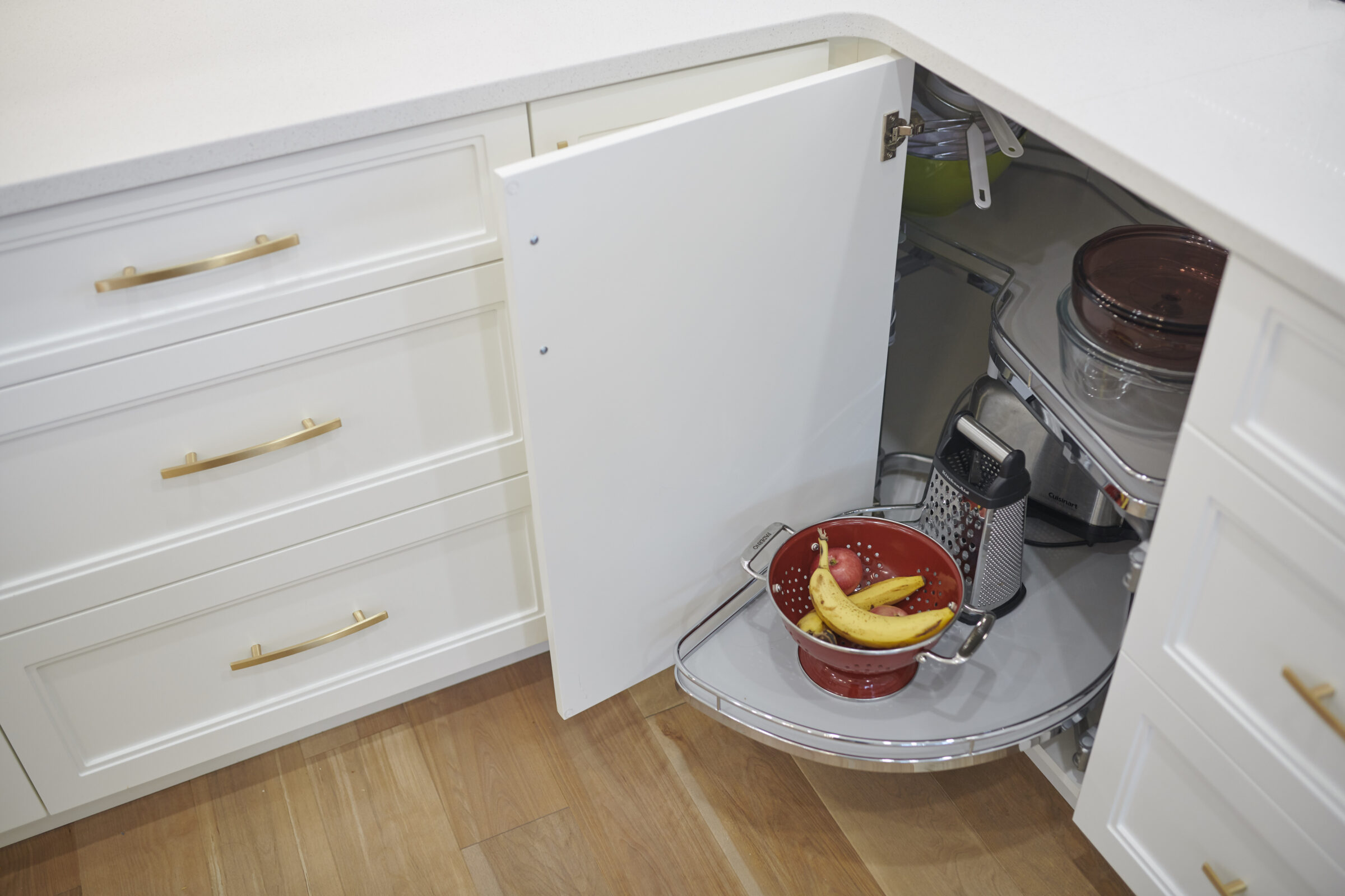 A corner kitchen cabinet with a white door is open, revealing a lazy Susan shelf system holding pots, a colander with fruit, and a grater.