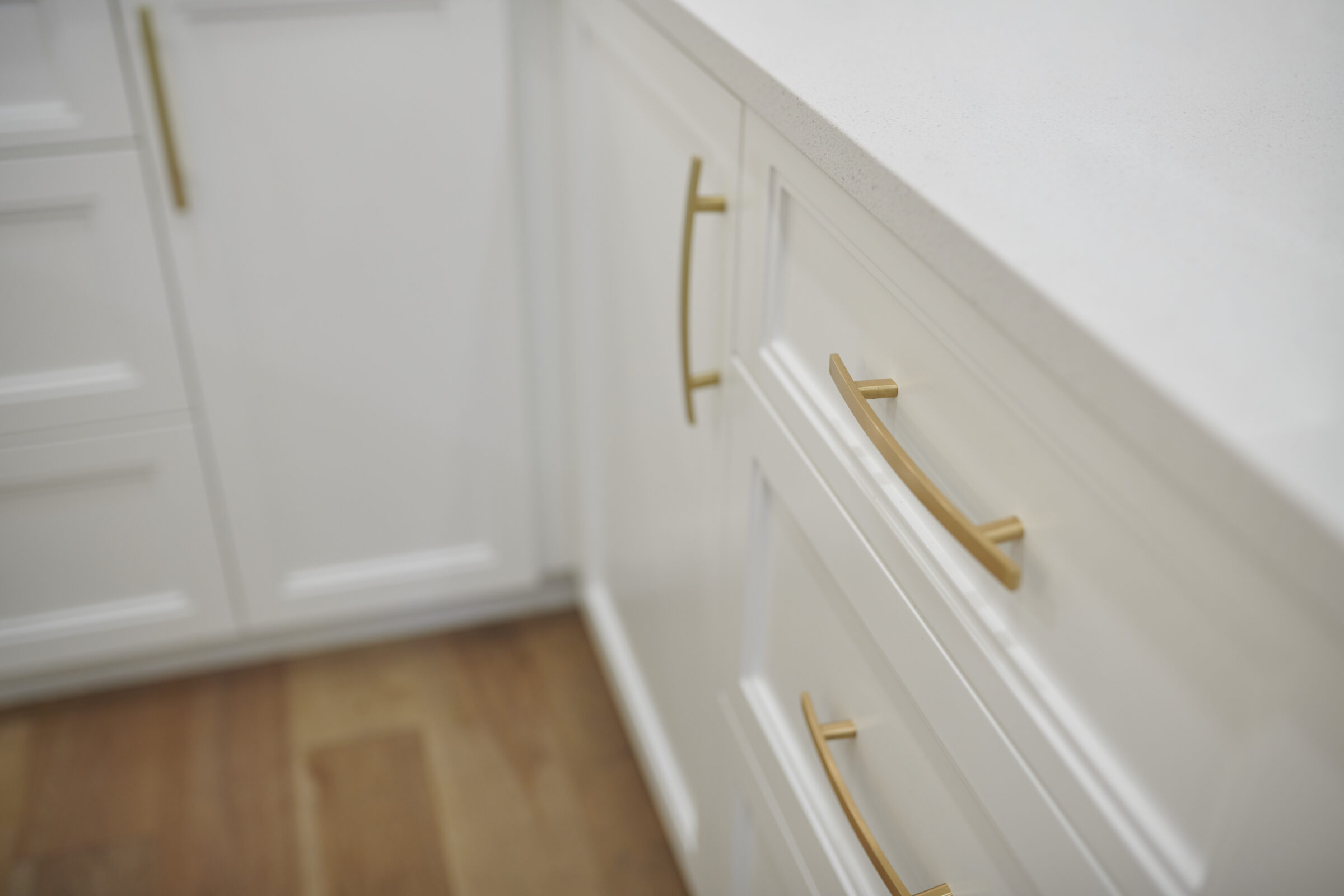 A modern kitchen with white cabinetry featuring sleek gold handles, a white countertop, and a warm wooden floor, all in soft focus.