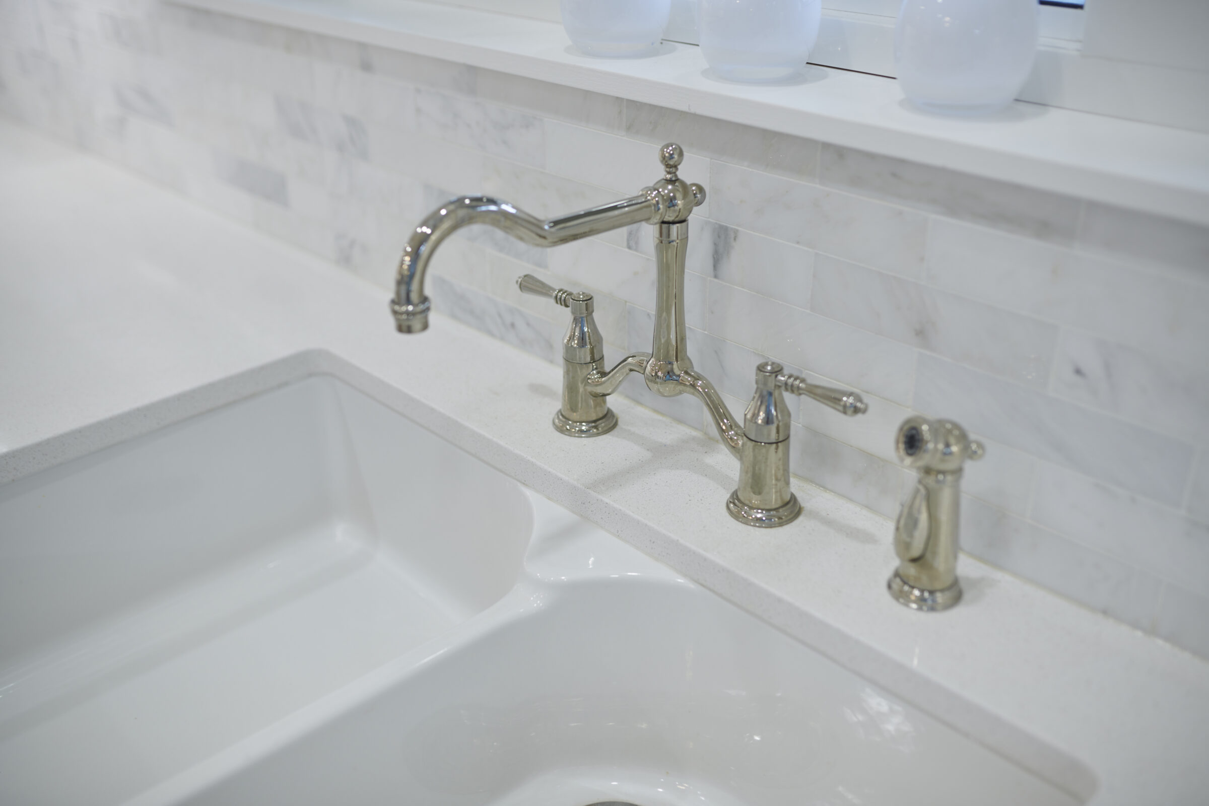 A modern bathroom sink with a white countertop, undermount basin, and sleek silver faucet set against a marble-like tiled backsplash. Two white vessels are visible above.