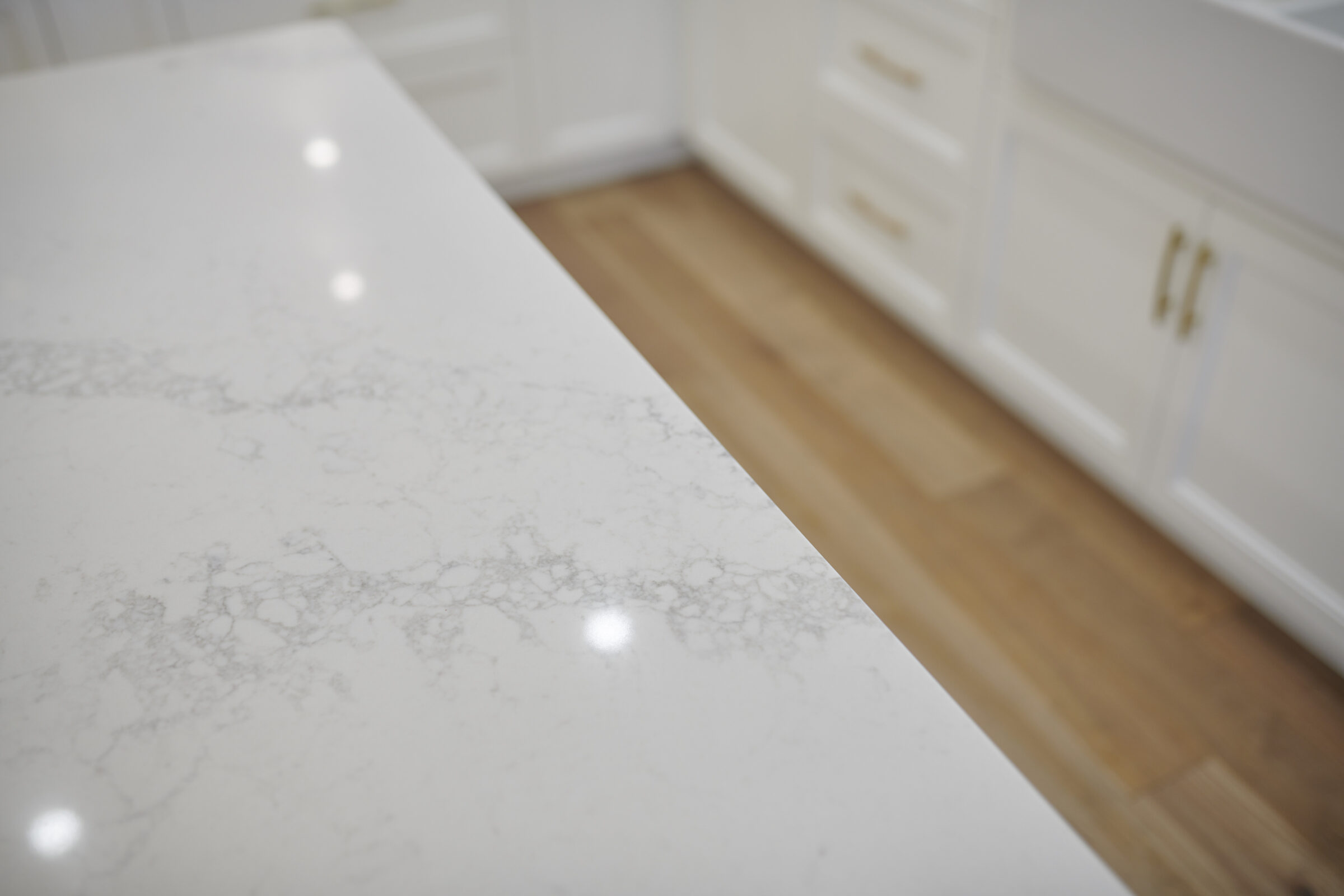 A close-up view of a white marble countertop with a reflective surface, showing light spots, adjacent to white cabinets and a wooden floor.
