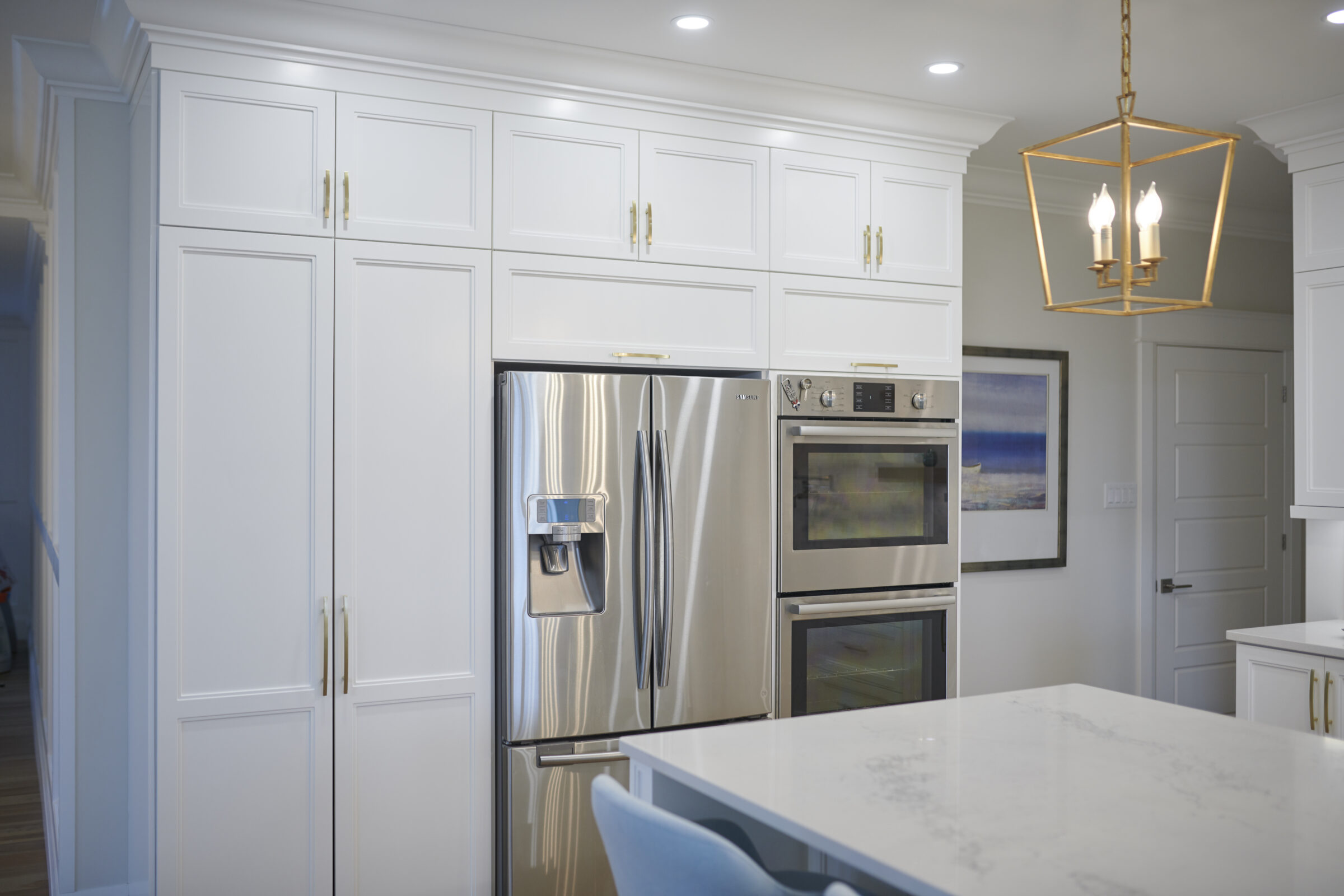 Modern kitchen with white cabinetry, stainless steel refrigerator, double oven, and pendant lighting. There's also a painting and a marble island countertop.
