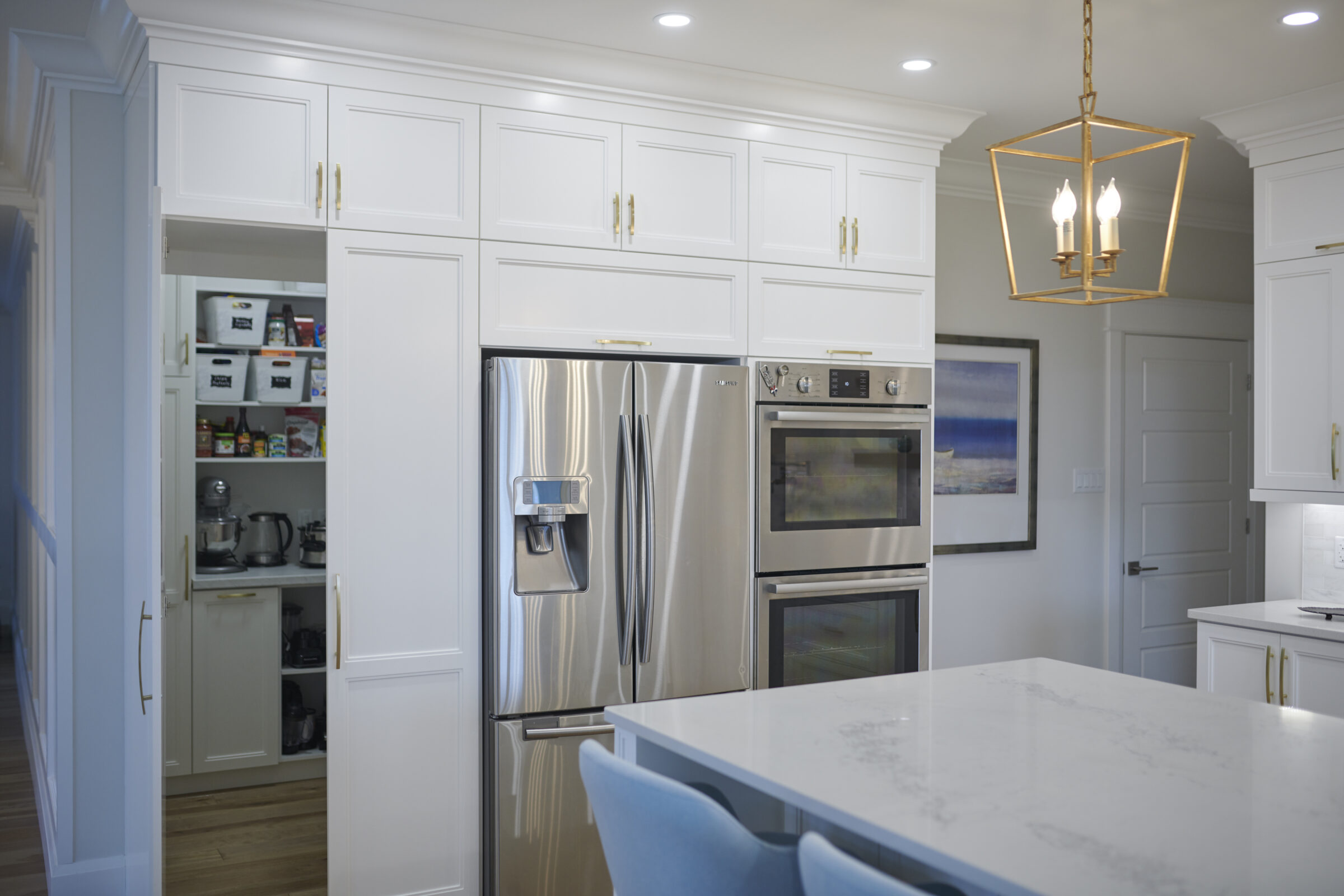 Modern kitchen interior with stainless steel appliances, white cabinets, marble countertop, blue chairs, geometric light fixture, and a partially open pantry door.