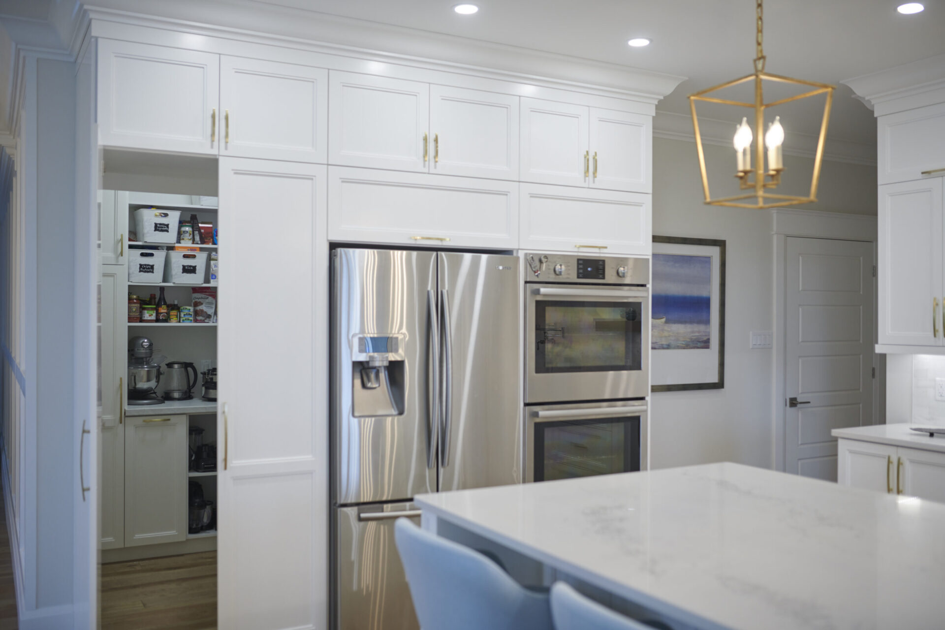A modern kitchen with white cabinetry, stainless steel appliances, a marble countertop island, blue chairs, and a geometric chandelier overhead.