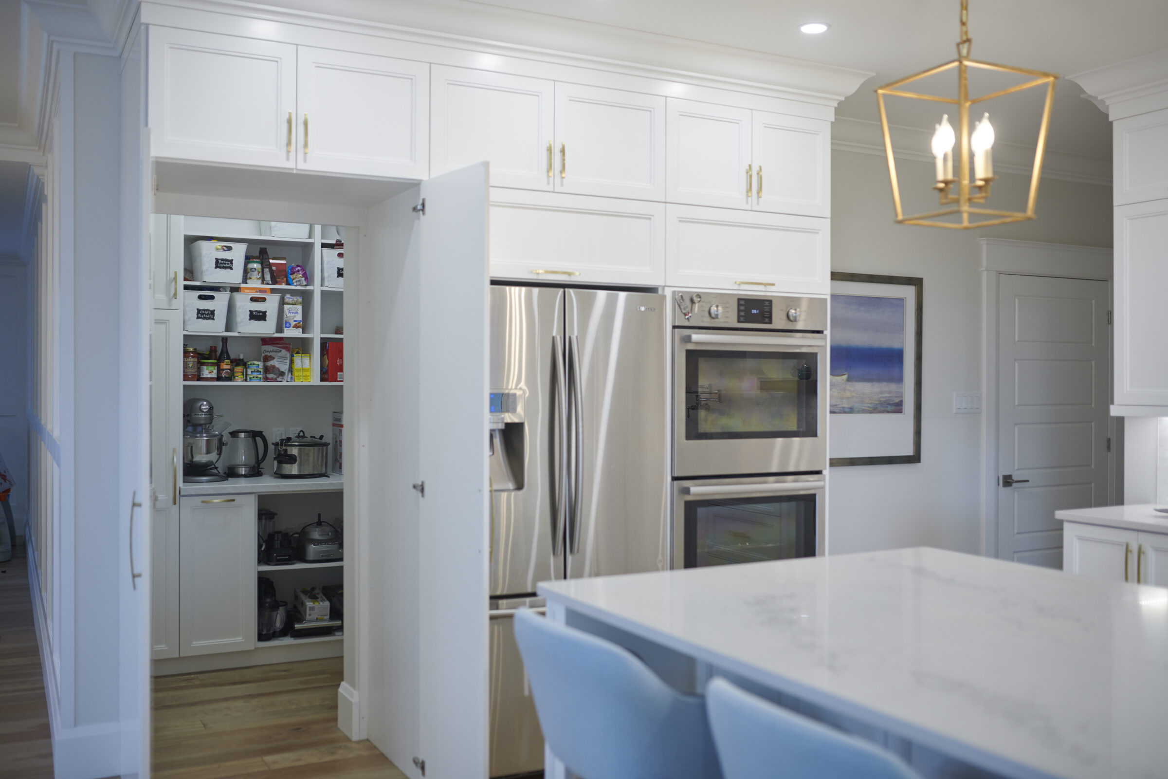 A modern kitchen with white cabinetry and stainless steel appliances. An open pantry reveals stocked shelves. A blue chair and pendant light add color accents.