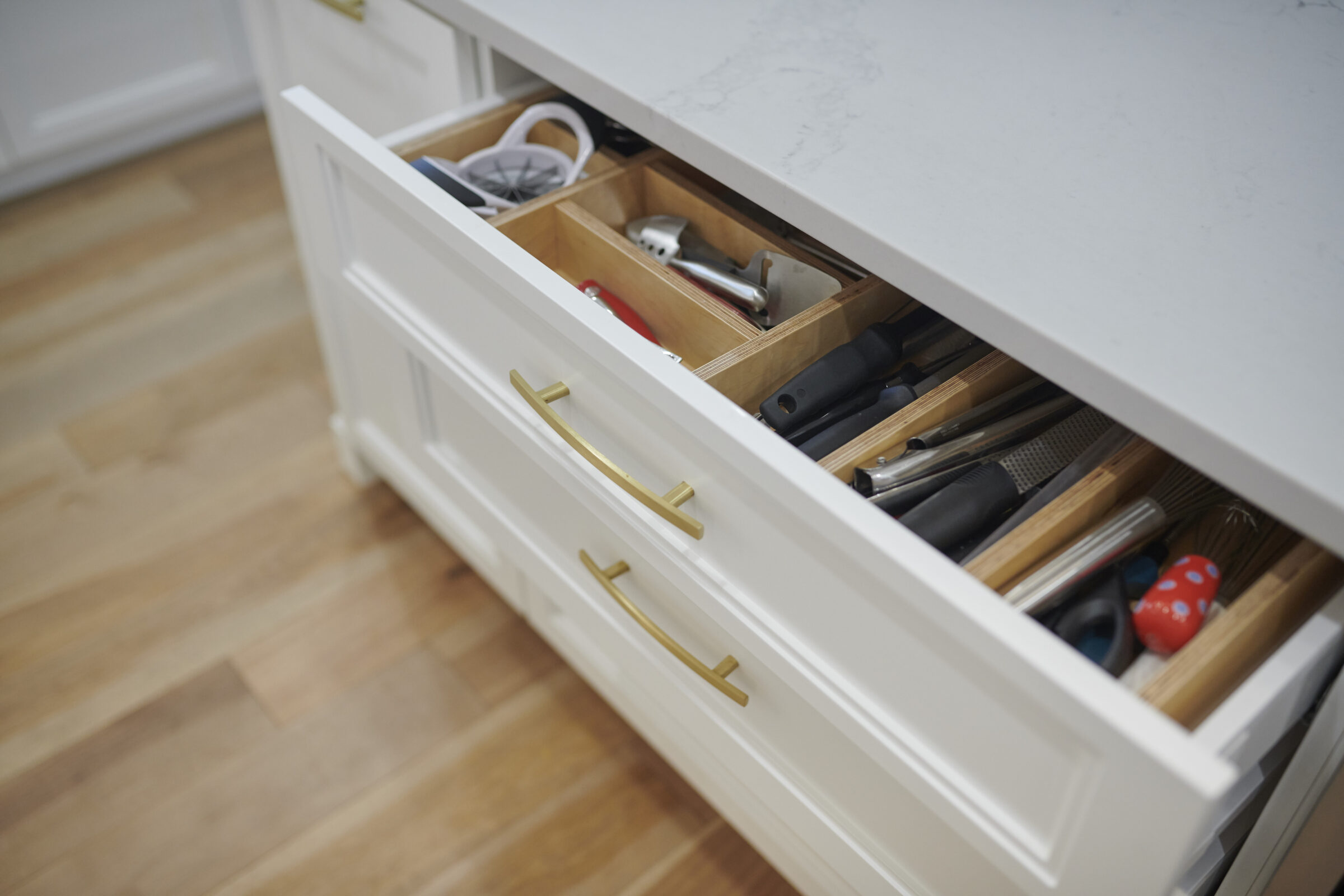 An open kitchen drawer revealing a variety of utensils; includes a knife block, whisks, and gadgets, with white cabinetry and gold handles.