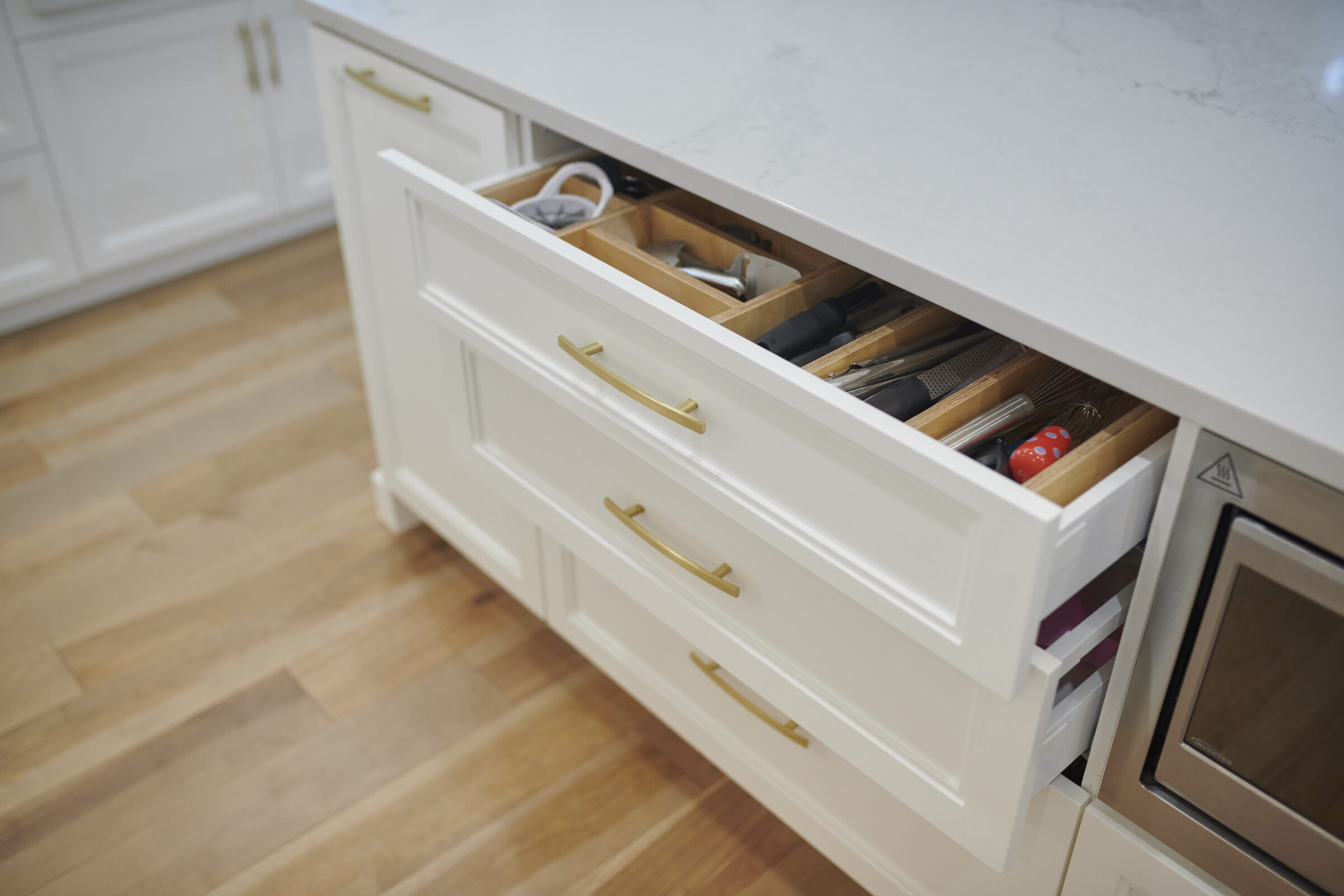 An open kitchen drawer reveals various utensils organized in compartments beside a closed white cabinet with gold handles and a built-in oven.