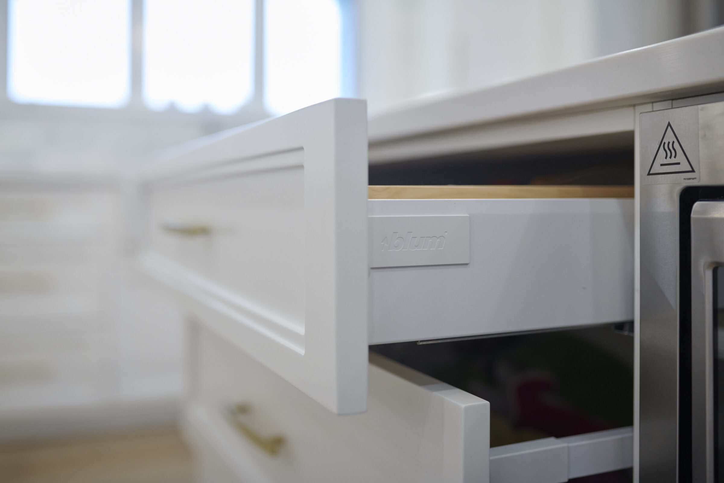 A close-up of a white kitchen drawer partially open, showing a modern design with a silver handle, next to a dishwasher with a caution symbol.