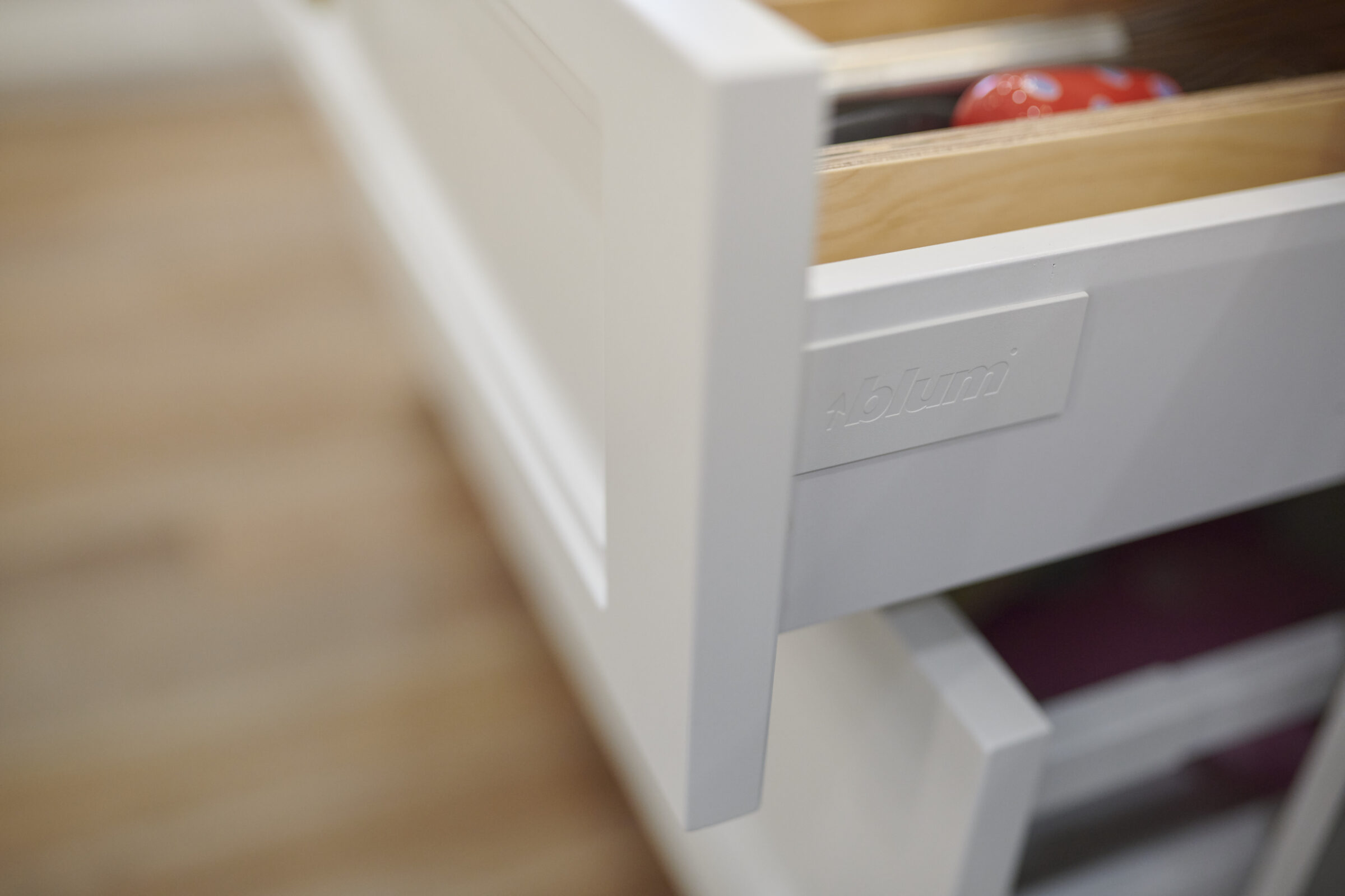 An open white drawer with a blurred view of kitchen items, with the brand "Blum" visible on the drawer side, in a clean, modern setting.