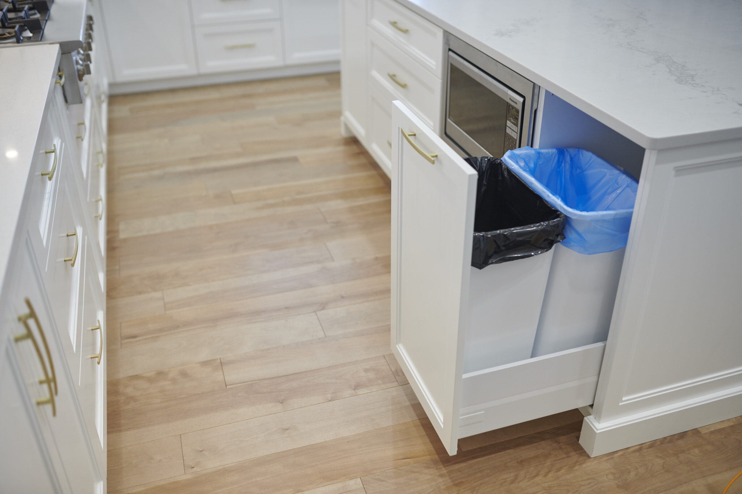 A modern kitchen with white cabinetry, gold handles, herringbone wood flooring, and a drawer bin with a blue recycling bag open.