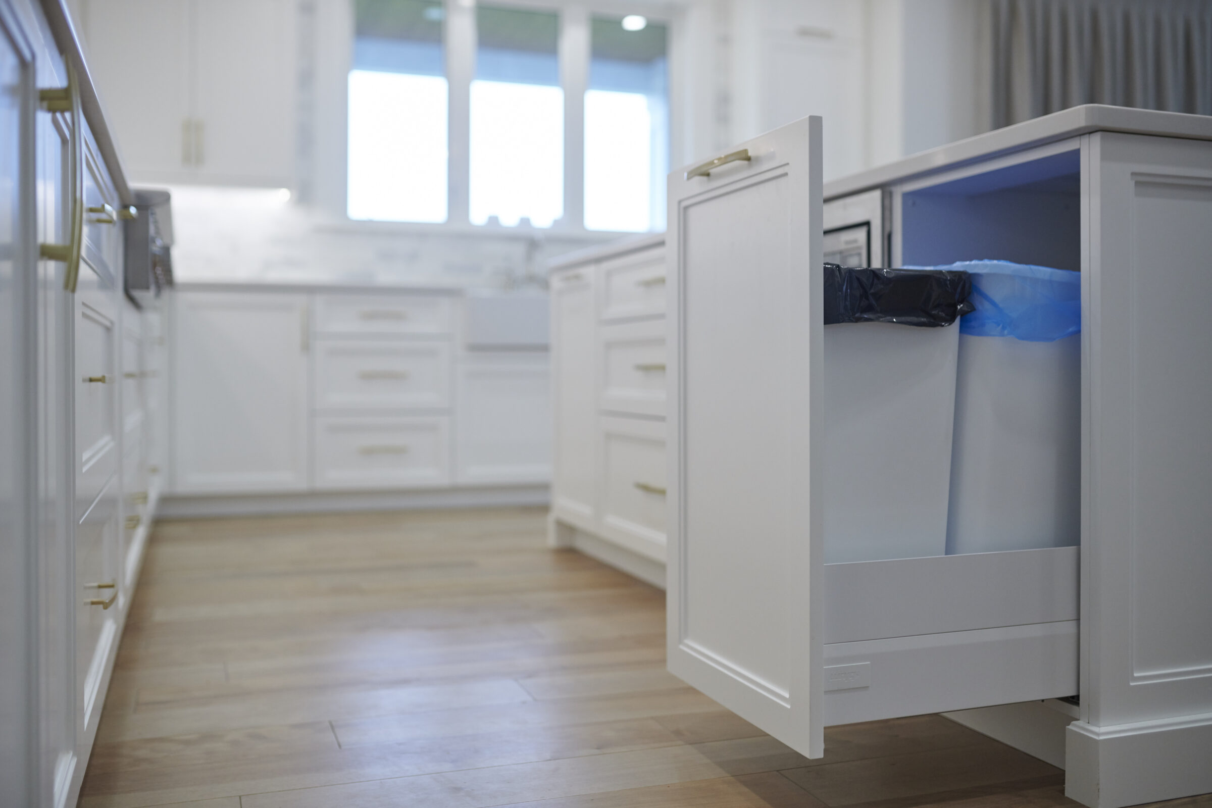 A modern kitchen with white cabinetry, brass handles, a built-in trash bin drawer open, and hardwood floors under soft daylight from windows.