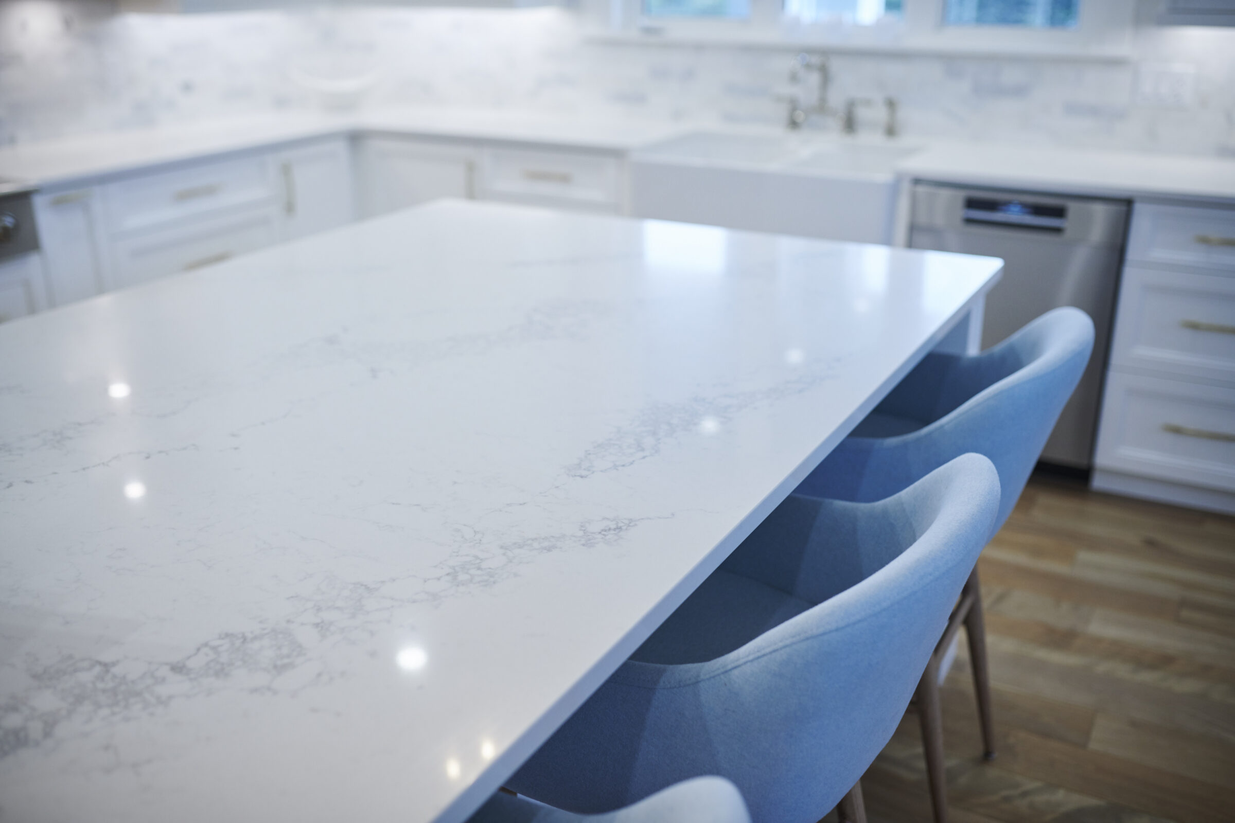 A contemporary kitchen featuring white cabinetry, marble countertops, and a central island with blue upholstered chairs. The flooring is light wooden.