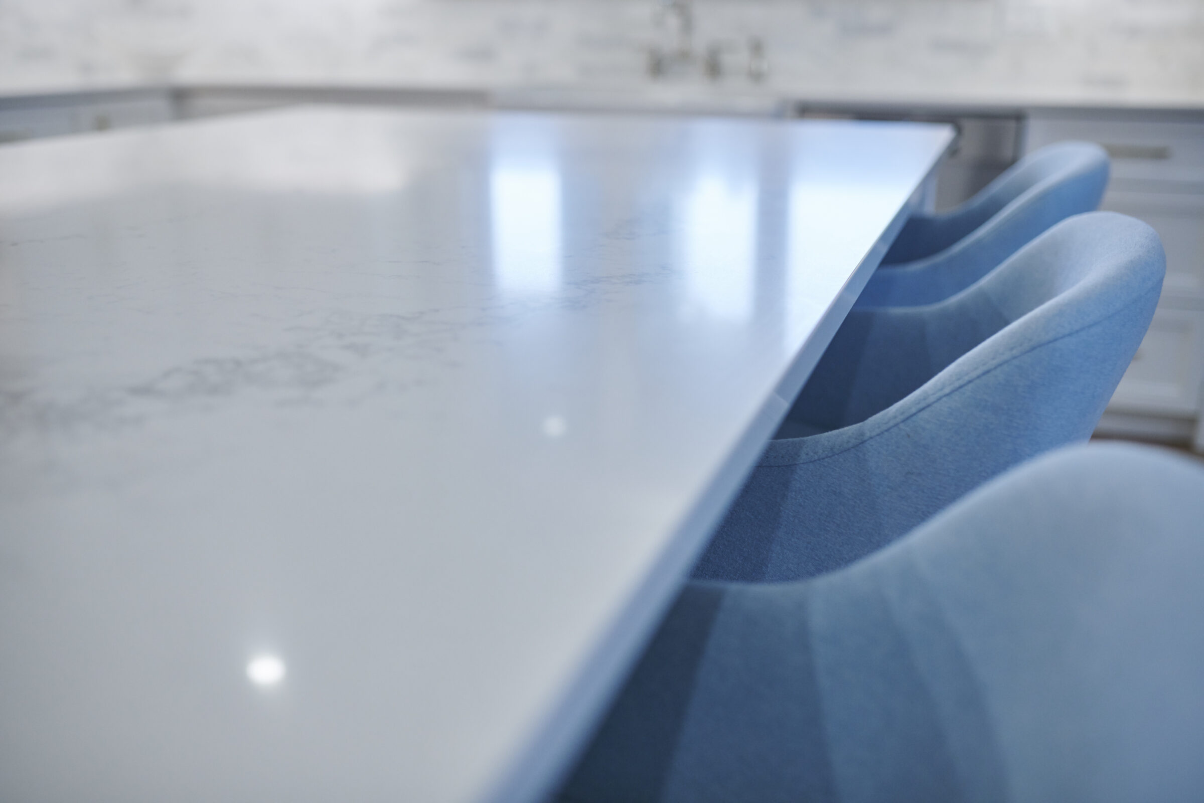 A modern kitchen interior with a glossy white countertop and blue upholstered chairs. The focus is soft, with natural light reflecting on surfaces.