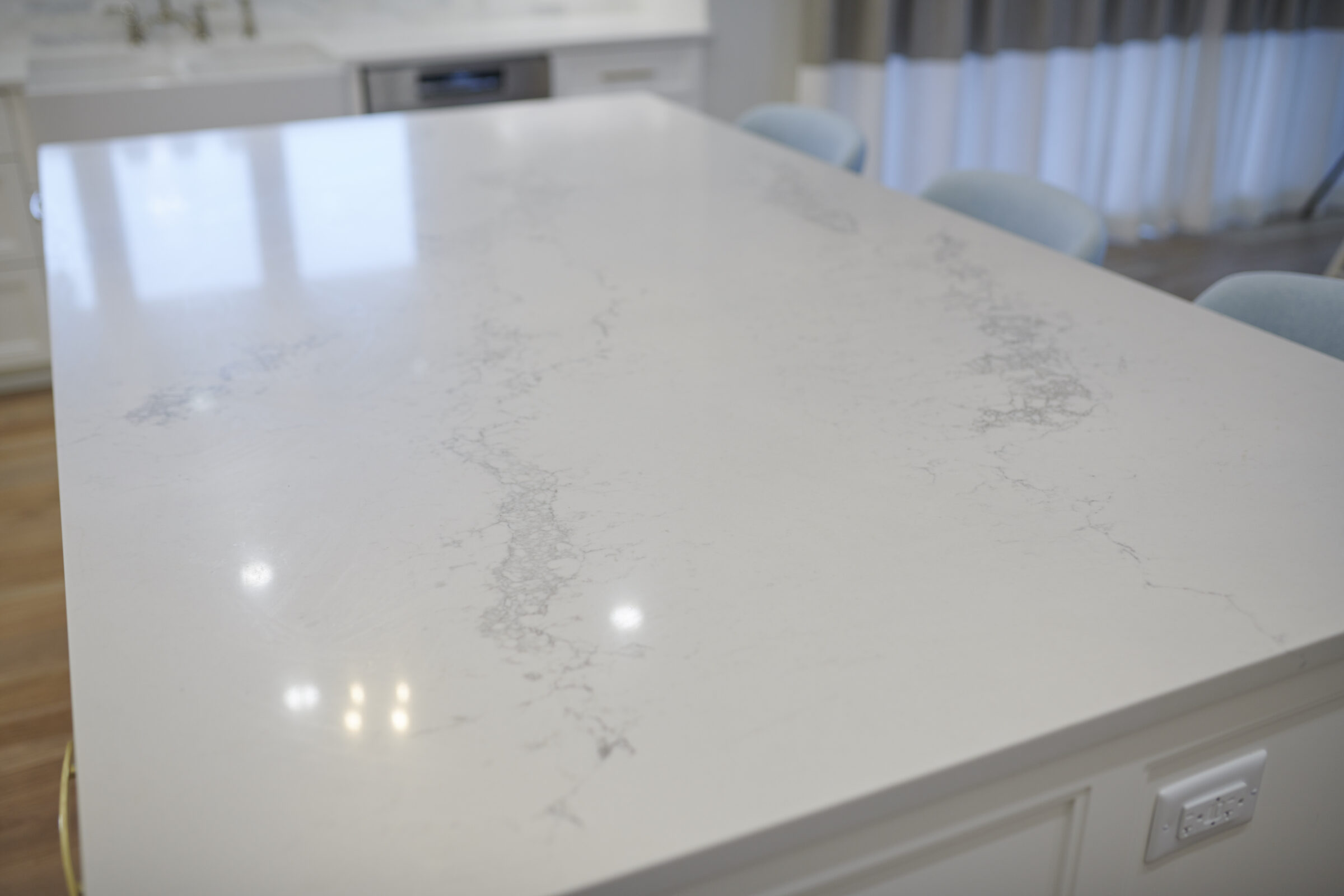 A modern kitchen featuring a large island with a white marble countertop, reflecting window light. Light blue chairs and white cabinets are visible.