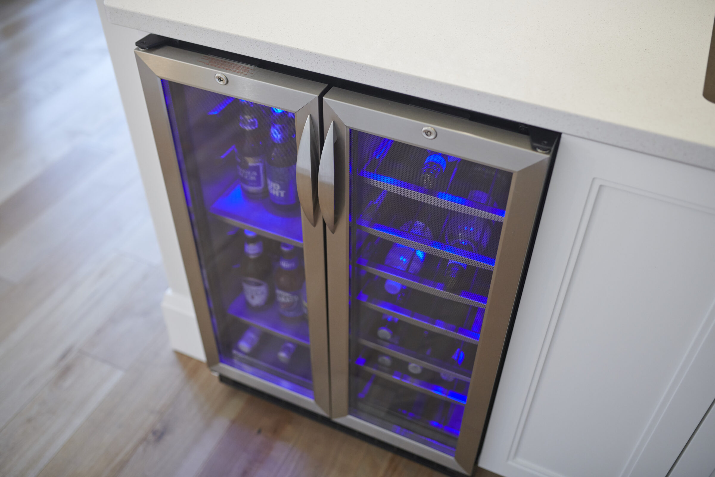 The image shows a modern, built-in dual zone wine cooler with glass doors, interior blue lighting, and shelves stocked with various bottles.