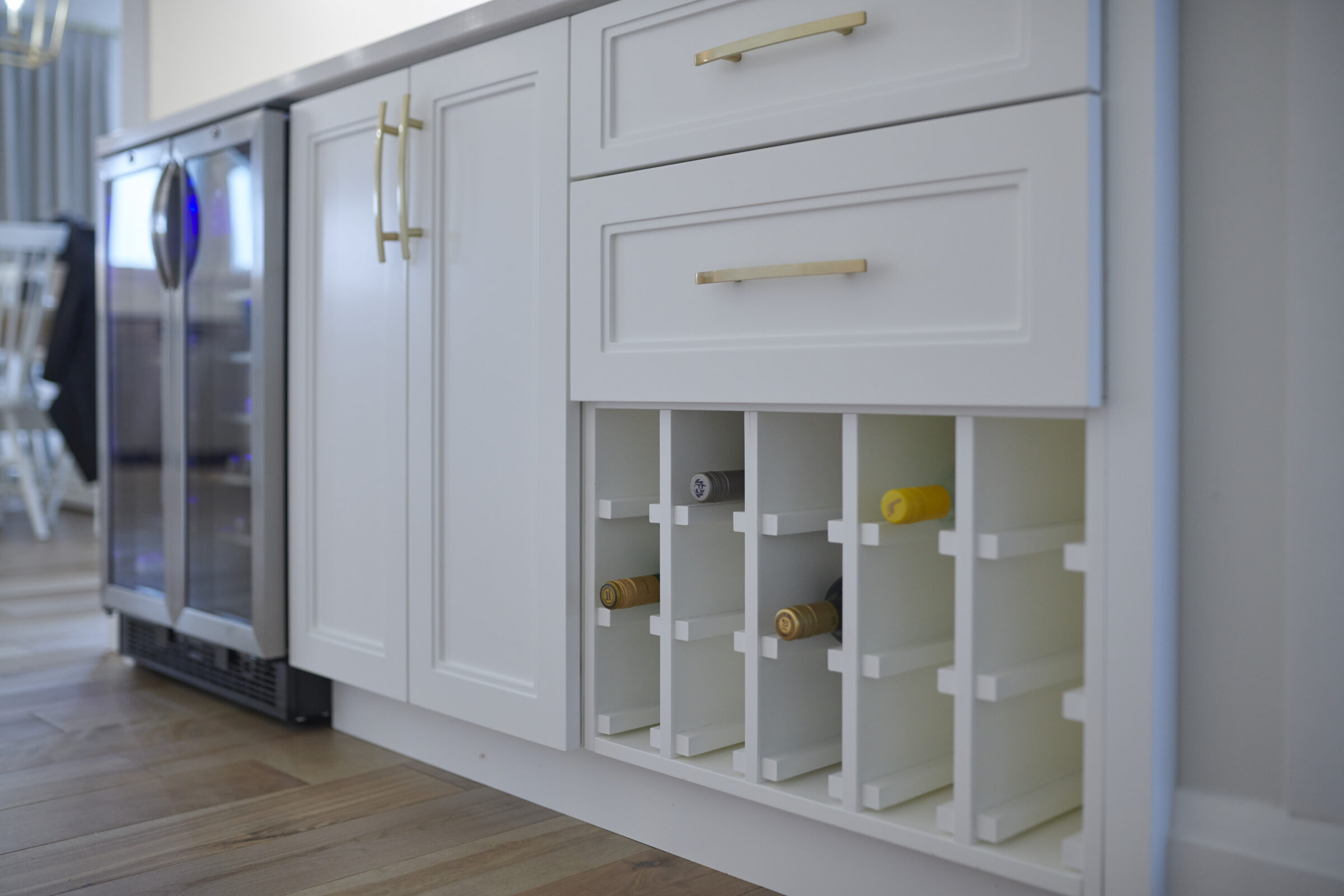 Modern kitchen interior featuring white cabinets with gold handles, drawers, a built-in wine rack with bottles, and a glimpse of a beverage cooler.