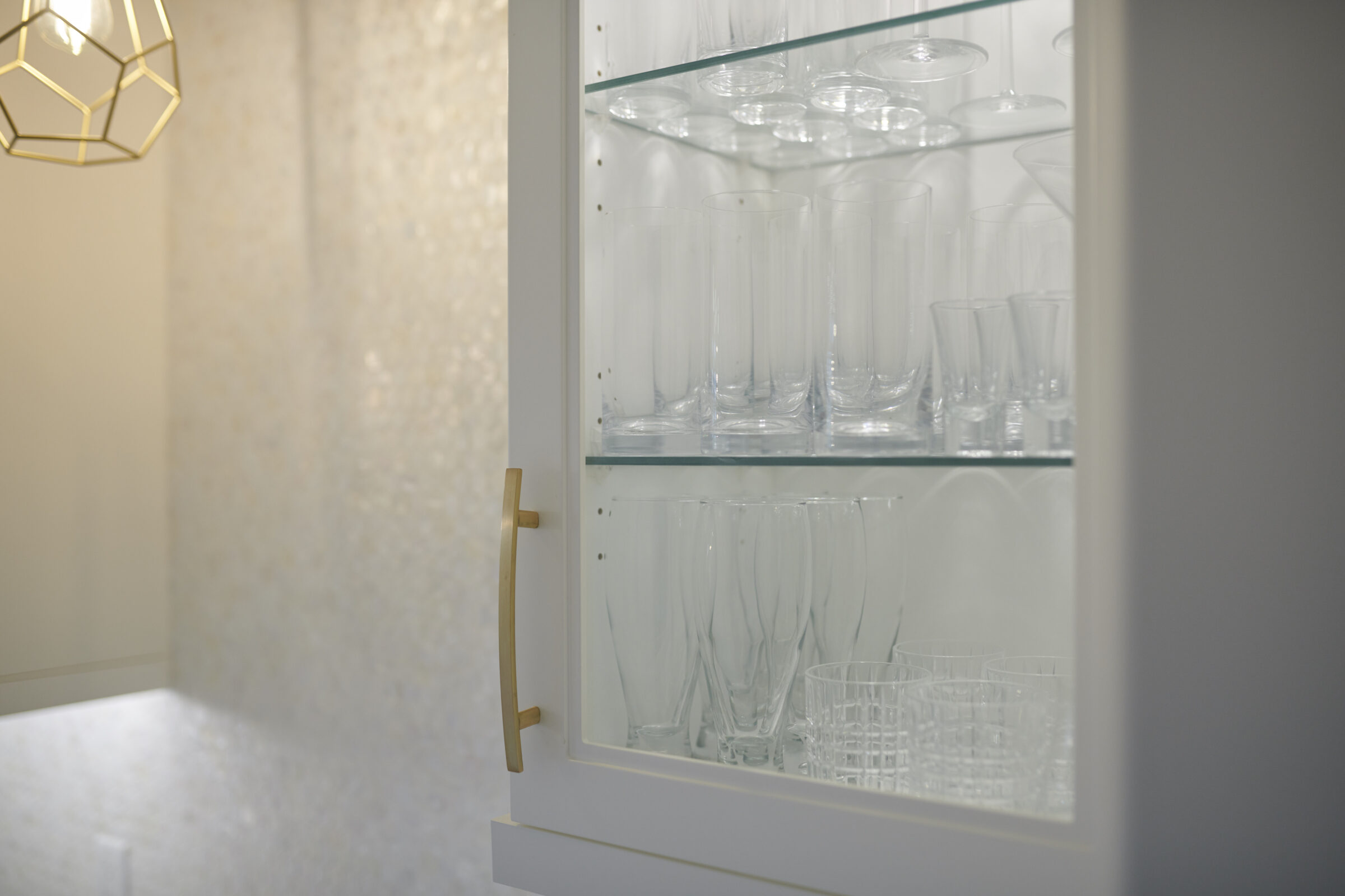 A glass-front cabinet displays various clear glasses on shelves, with a geometric light fixture visible in the softly lit, neutral-toned background.