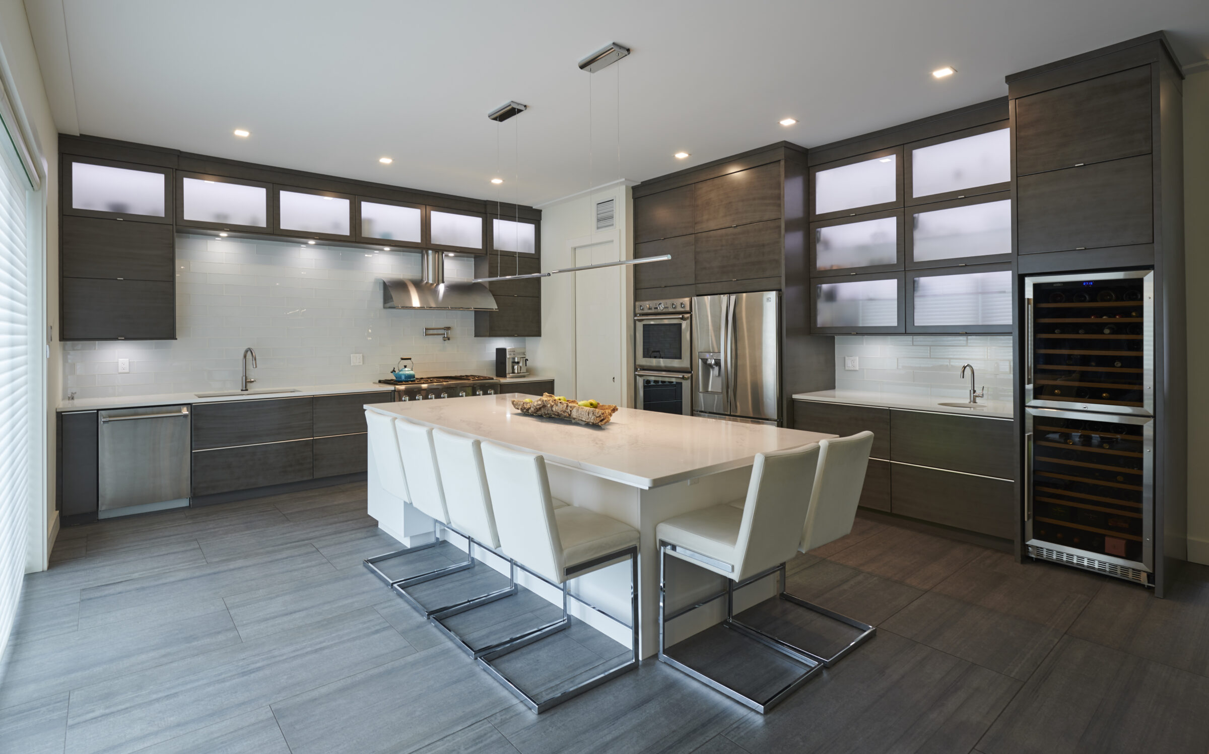 The image shows a modern kitchen with sleek, dark cabinetry, stainless steel appliances, a large island with white chairs, and a wine fridge.