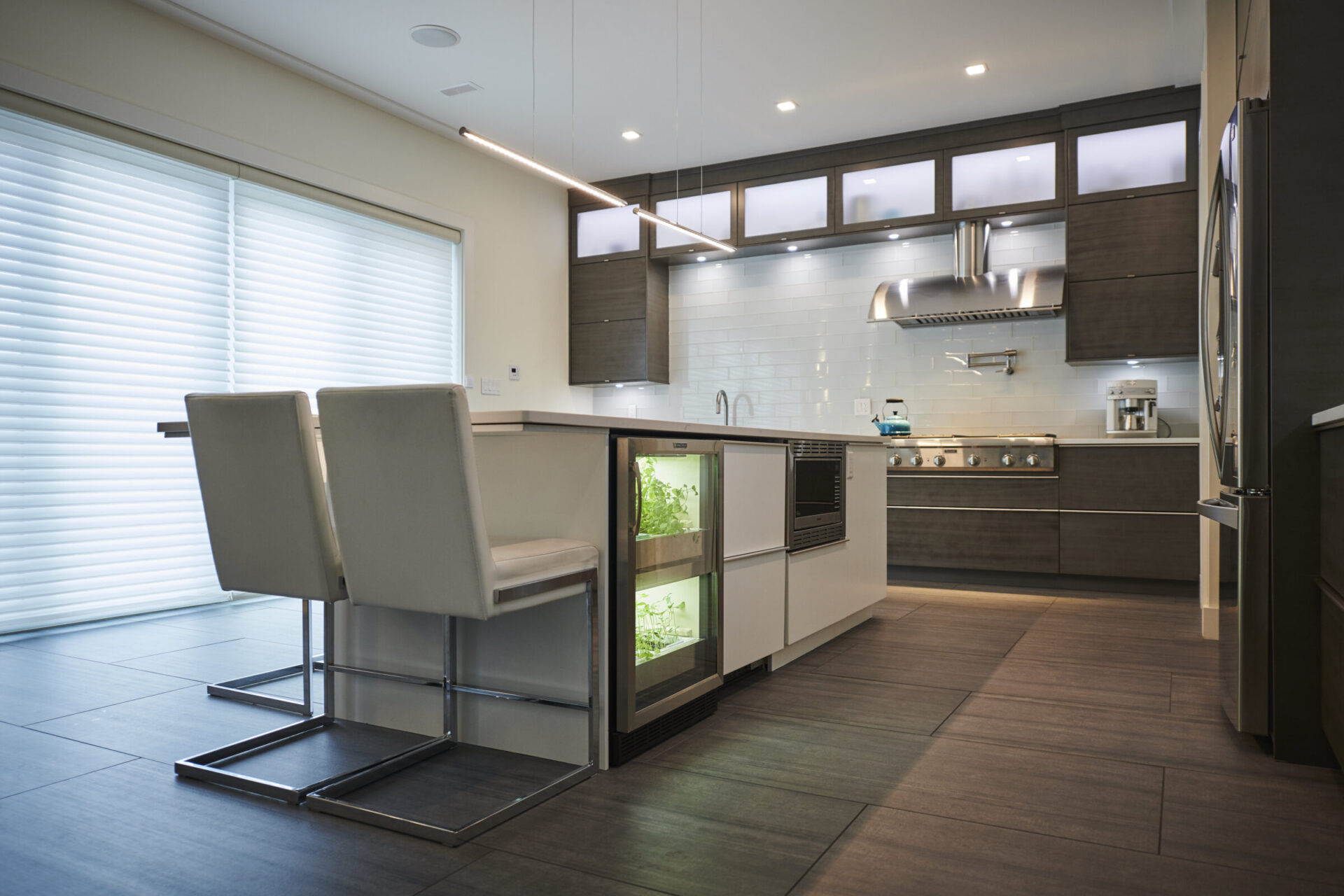Modern kitchen interior with dark wood flooring, white countertops, stainless steel appliances, bar stools, and abundant natural light filtering through blinds.