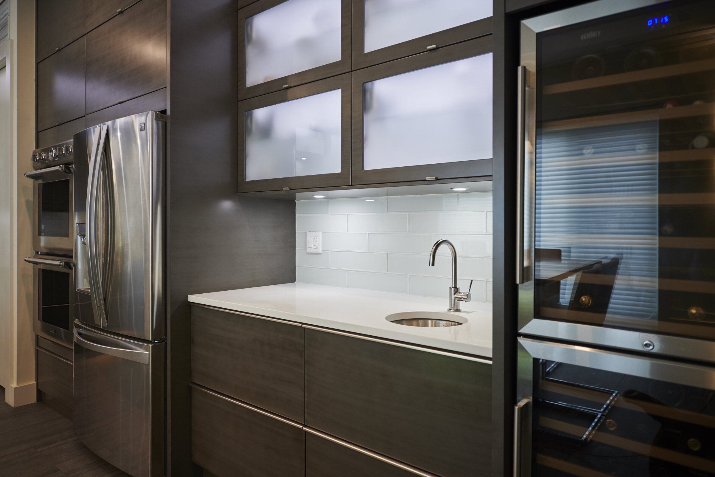 Modern kitchen interior with stainless steel appliances, wooden cabinets, white countertop, built-in oven and microwave, and a wine fridge.