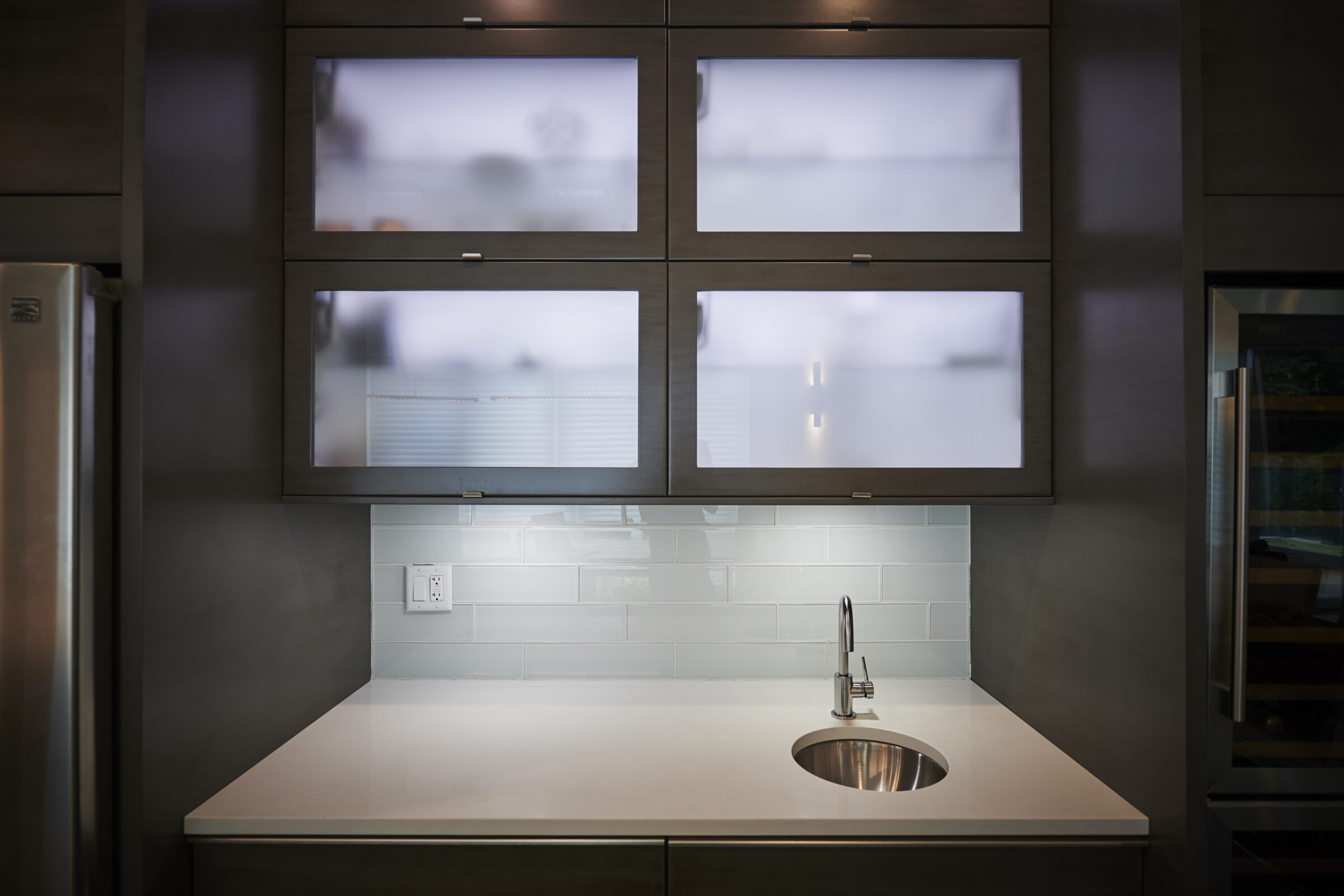 This is a modern kitchen with frosted glass cabinet doors, a stainless steel sink, white backsplash tiles, and a refrigerator to the right.