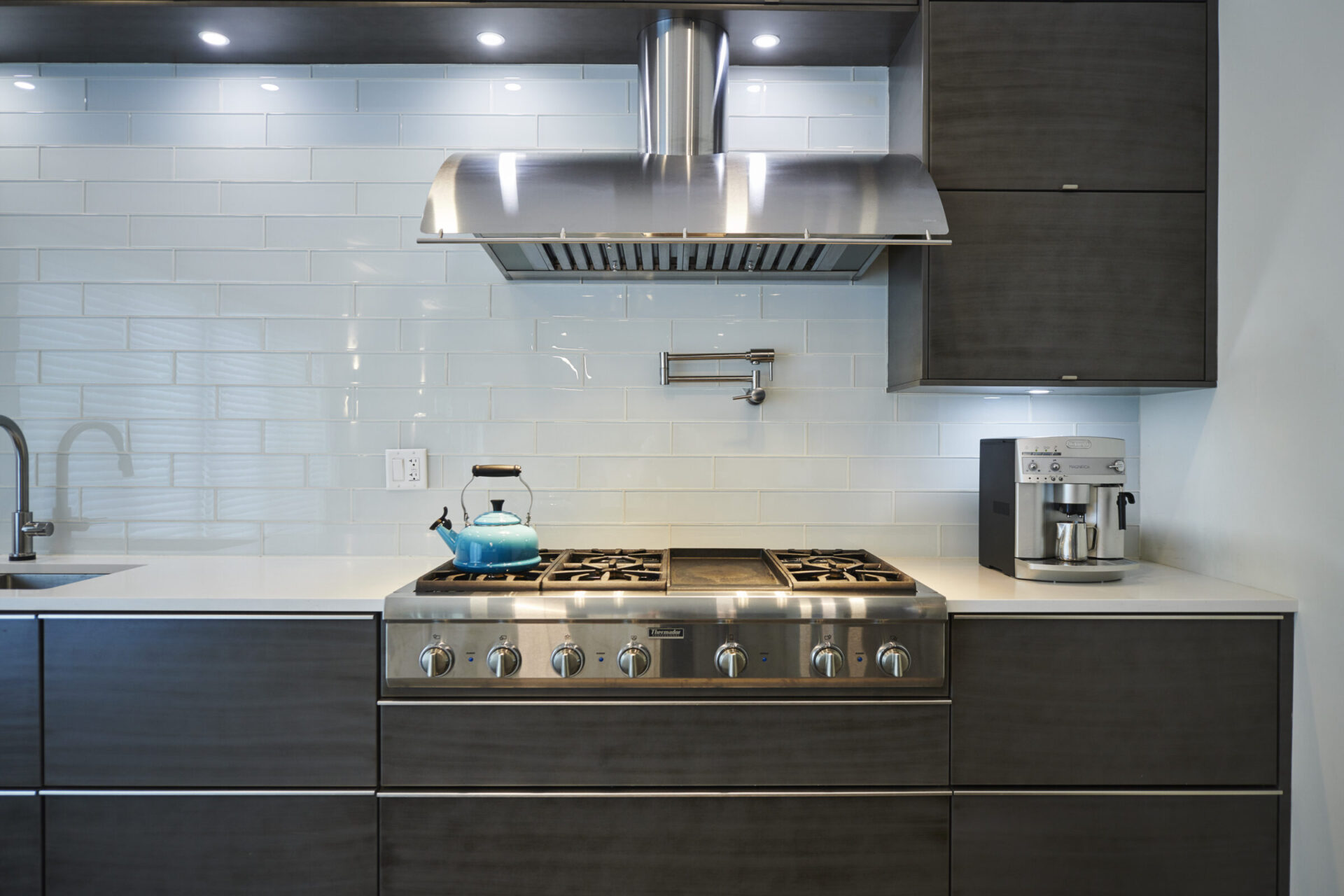 Modern kitchen interior with stainless steel gas stove, blue kettle, large range hood, dark cabinetry, white countertops, and subway tile backsplash.
