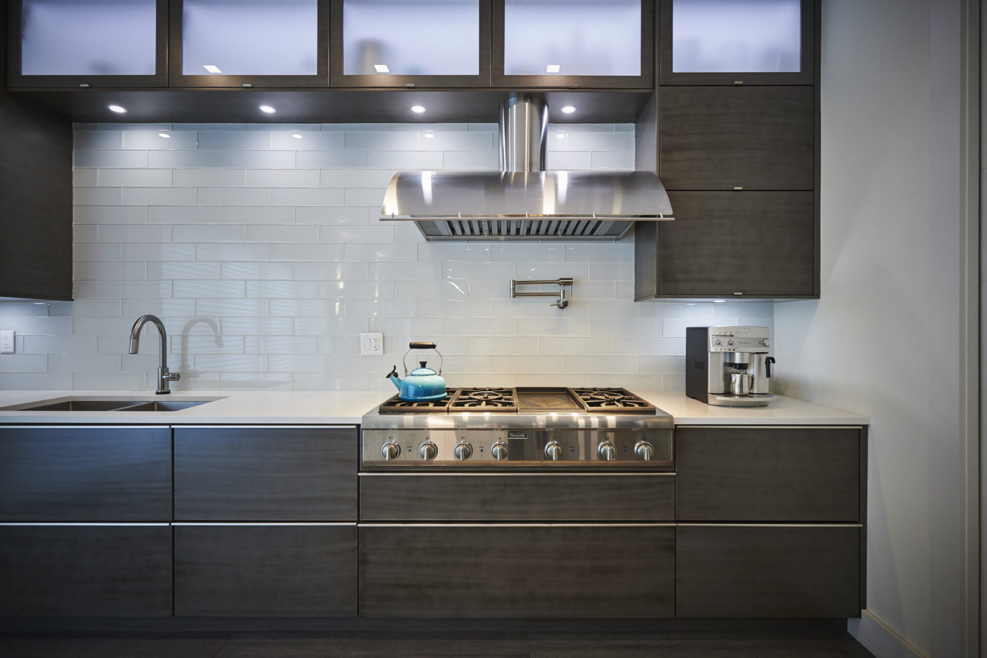 A modern kitchen featuring stainless steel appliances, dark cabinetry, a gas stove, and a teal kettle. White subway tile backsplash and under-cabinet lighting accent the space.