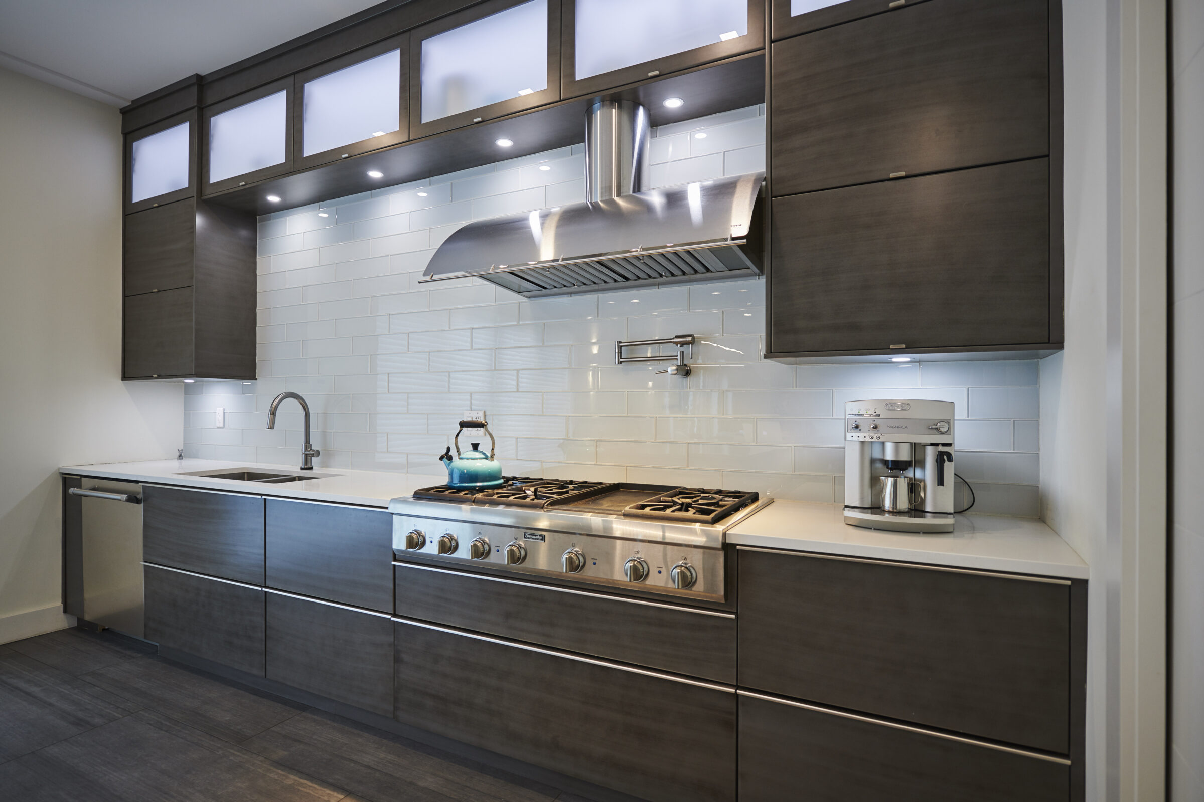 A modern kitchen with stainless steel appliances, dark wood cabinetry, white countertops, subway tile backsplash, and hardwood flooring, showing a clean, sleek design.