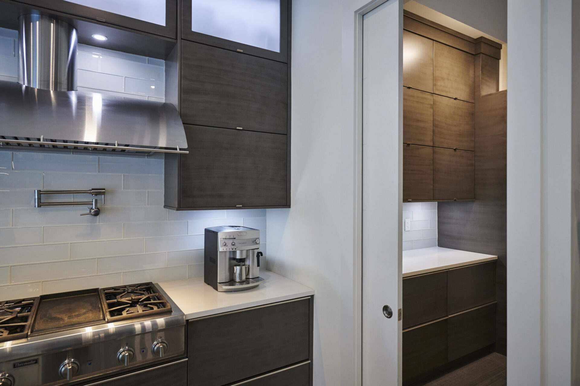 Modern kitchen with stainless steel gas stove, range hood, espresso machine, blue backsplash tiles, and dark wood cabinets opening to a small hallway.