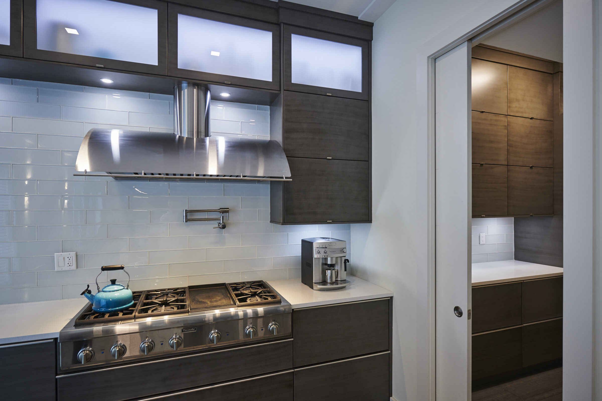 Modern kitchen with stainless steel gas stove, blue tea kettle, espresso machine, dark wood cabinets, frosted glass panels, and white subway tile backsplash.