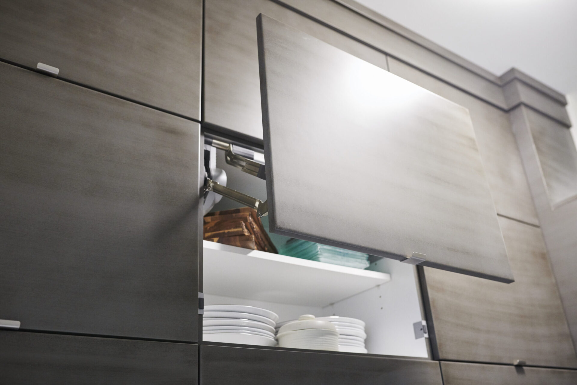 This image shows a modern kitchen cabinet with a lift-up door mechanism revealing stacked white dishes and bowls inside. The design is sleek and contemporary.