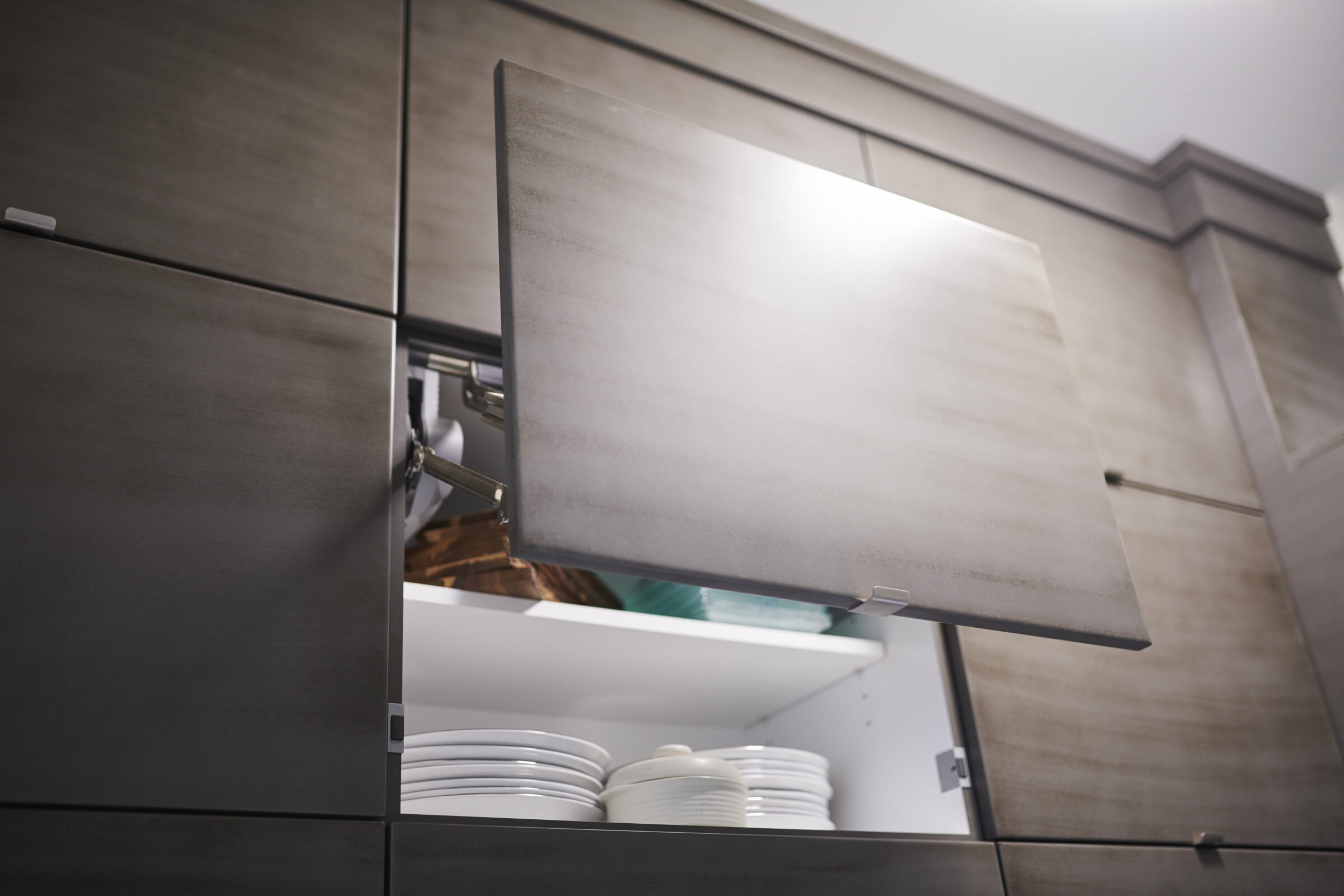 A modern kitchen cabinet is open, revealing stacked plates within. The cabinet features a metallic finish with a handle and hinges visible.