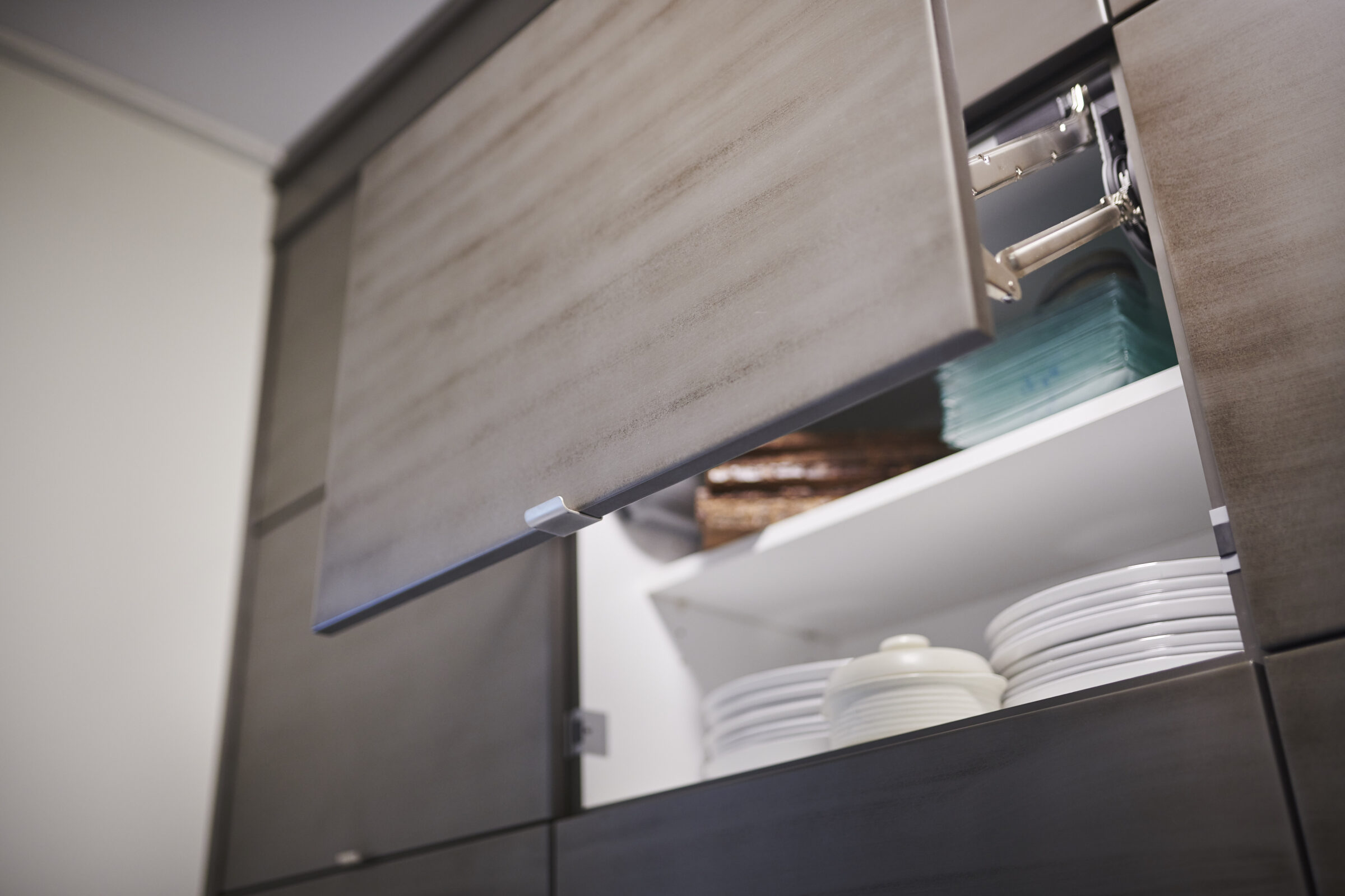 Modern kitchen cabinet with an open upper door, revealing neatly stacked plates and glassware, featuring a minimalist design with a light wood finish.
