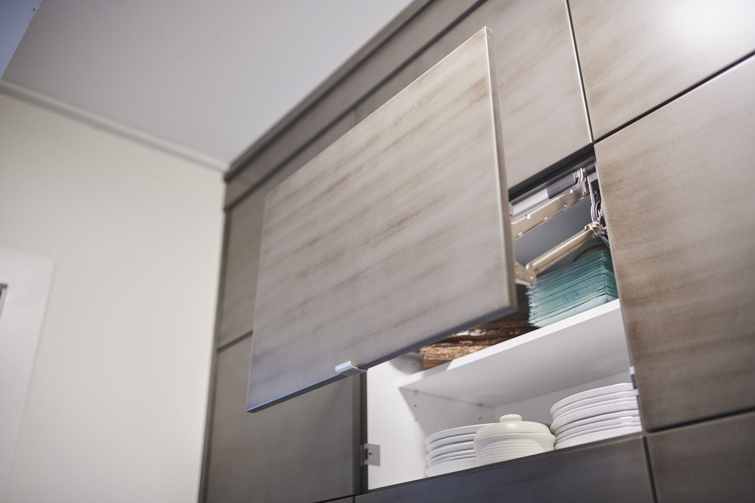 This image shows an open modern kitchen cabinet with a collection of white plates and glass shelves. The design is sleek and minimalist.