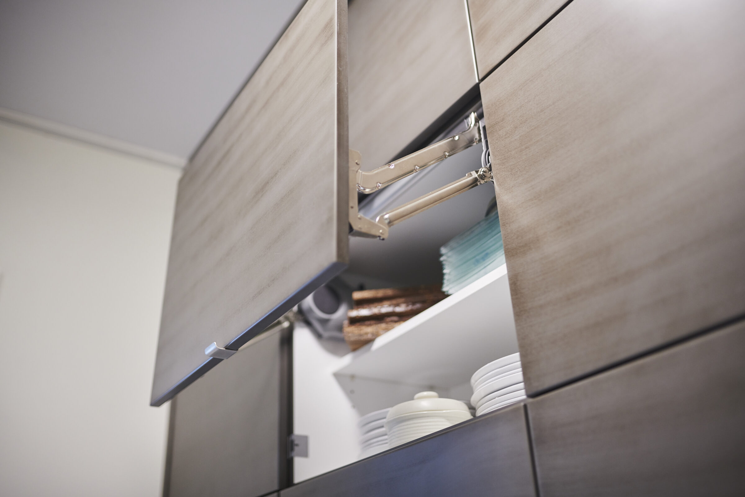Modern kitchen cabinet with an open door, showcasing plates and glassware inside. The cabinet features a metallic handle and hinge mechanism.