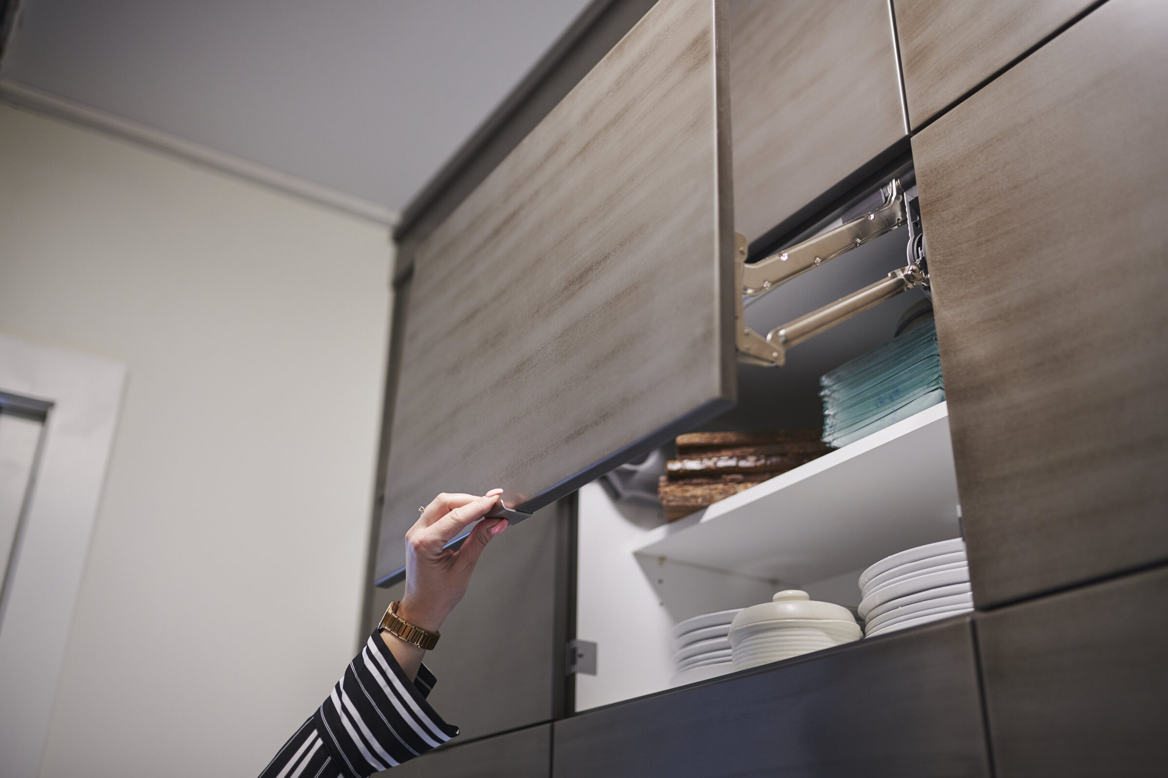 A person is reaching for a modern kitchen cabinet with a lifted door, revealing neat stacks of plates and kitchen items inside.
