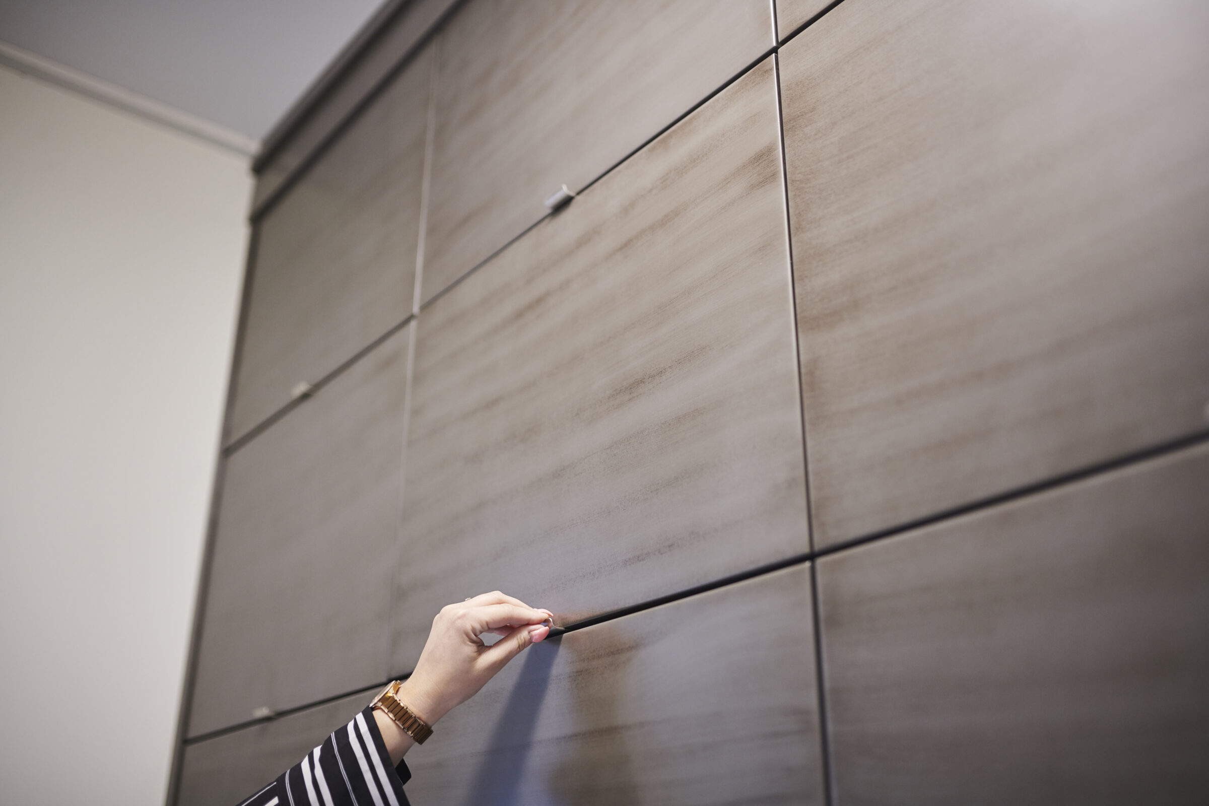 A person's hand is opening a wooden cabinet drawer. They are wearing a striped shirt and a bracelet. The room has a modern design.