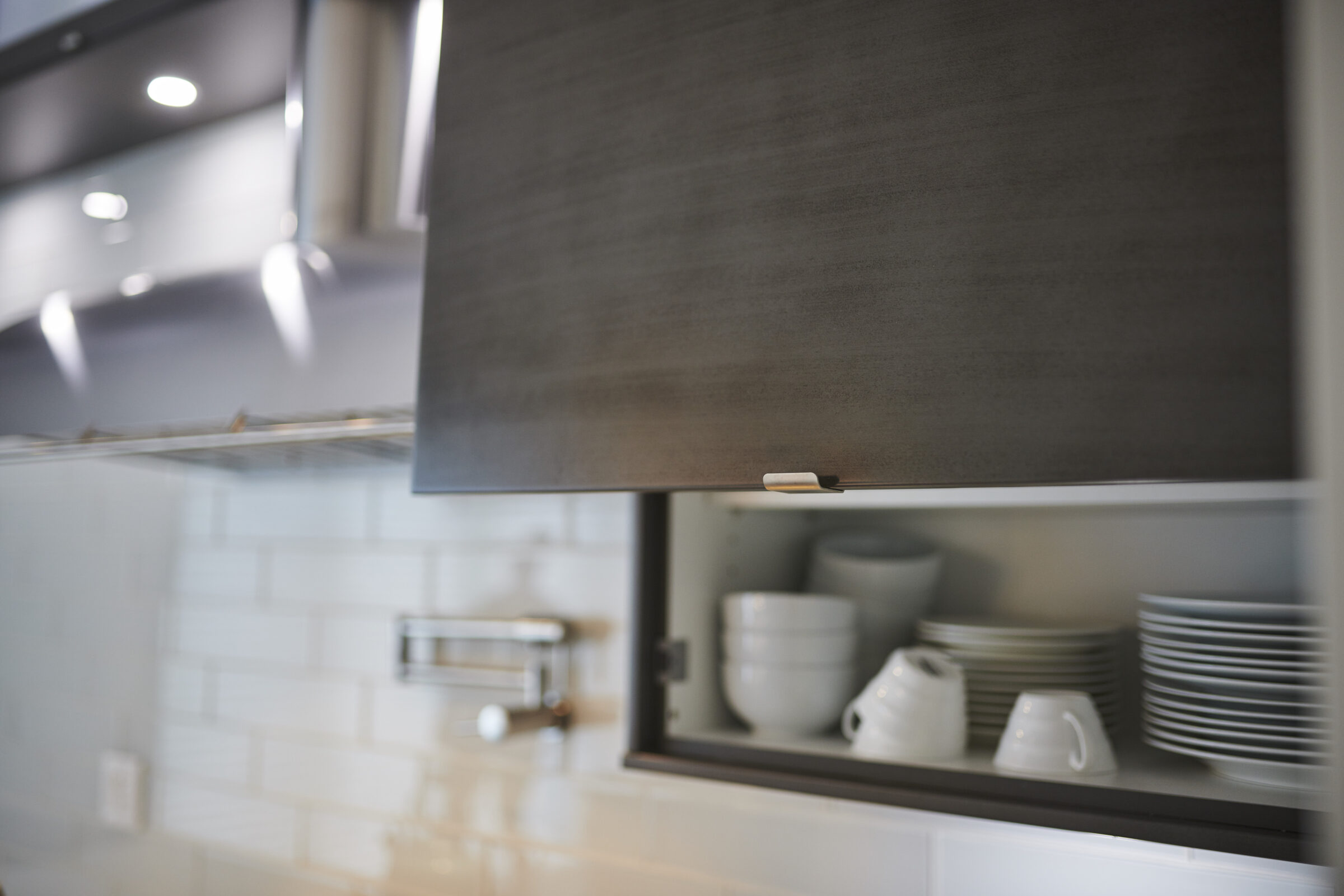 This is a modern kitchen interior showing an open cabinet with white dishes and bowls, under a gray roller shade, with subdued lighting.