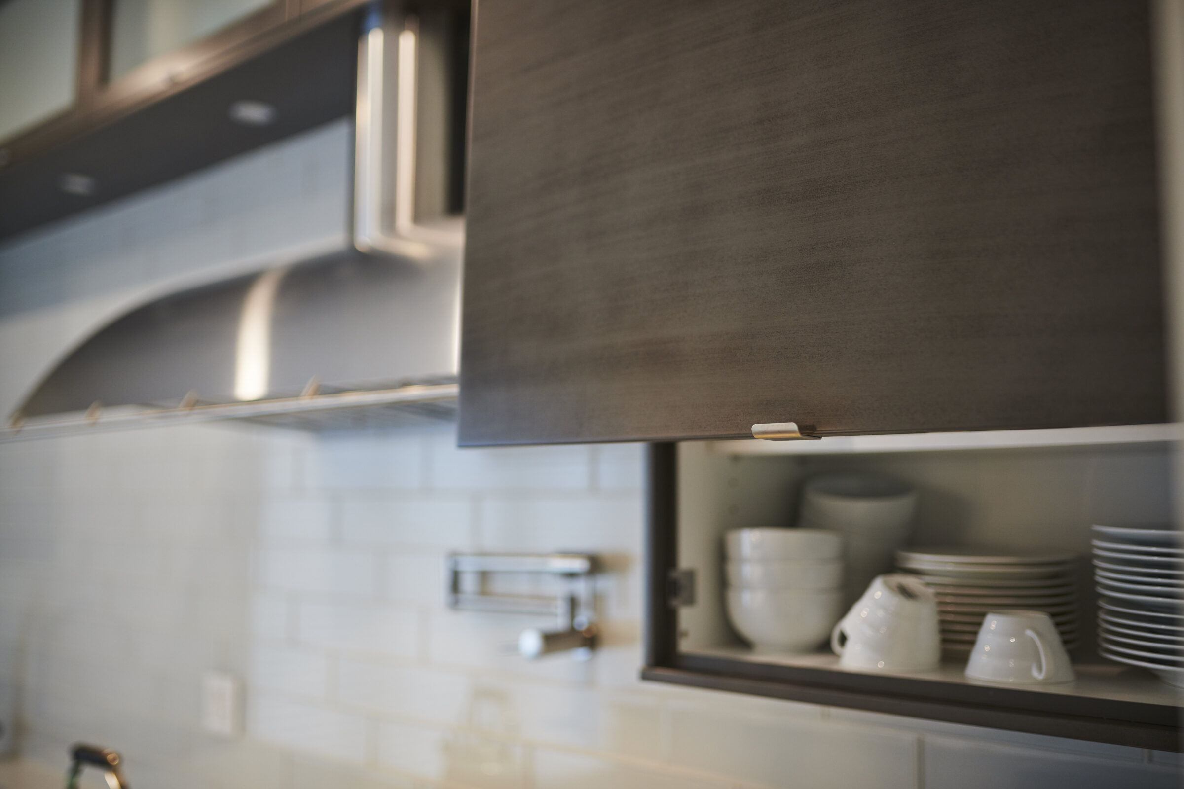 Modern kitchen with open wooden cabinet displaying white dishes. Stainless steel range hood, subway tile backsplash, and blurred foreground create a clean aesthetic.