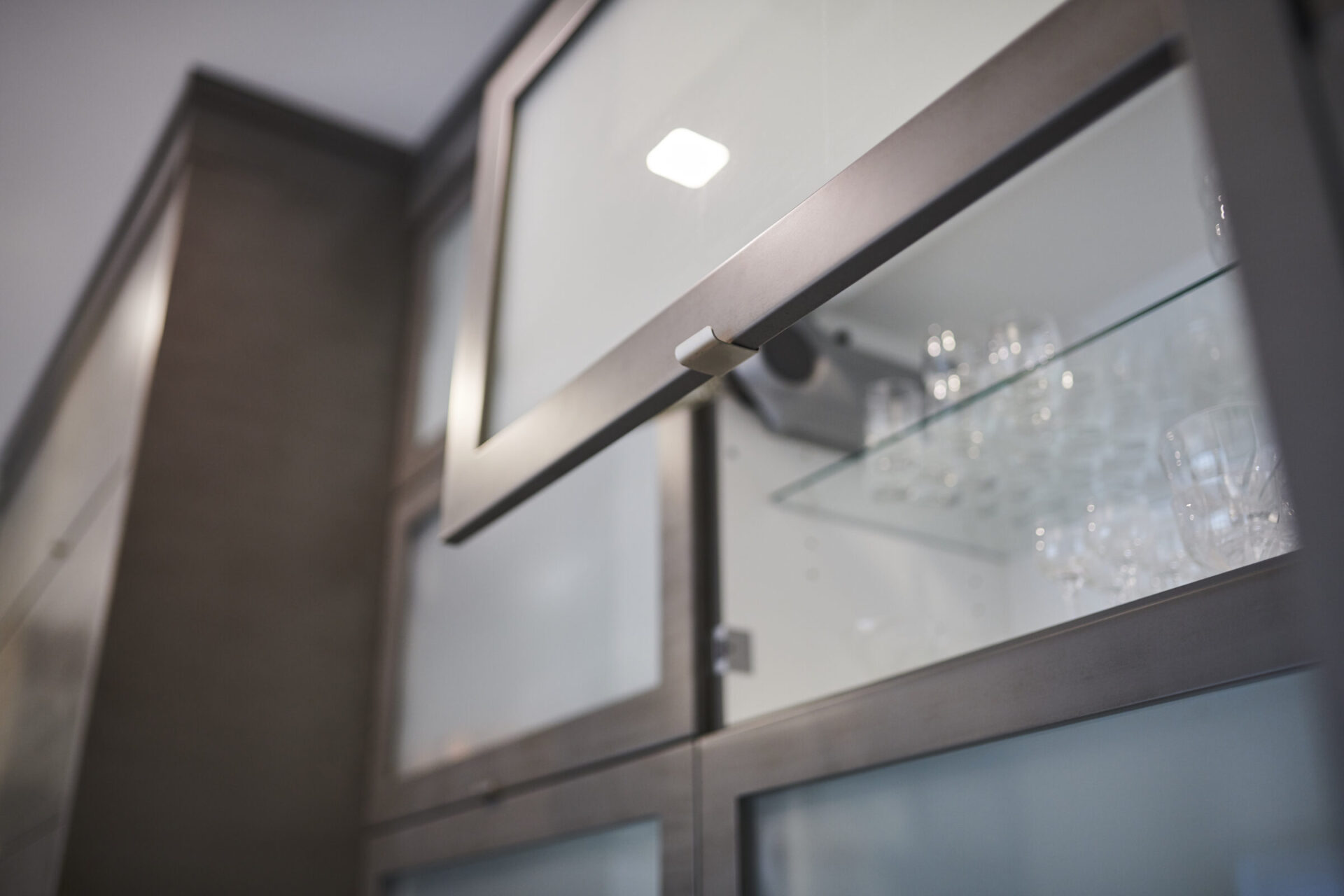 A close-up view of a modern kitchen cabinet with frosted glass doors, highlighting the sleek metal handle with wine glasses visible inside.