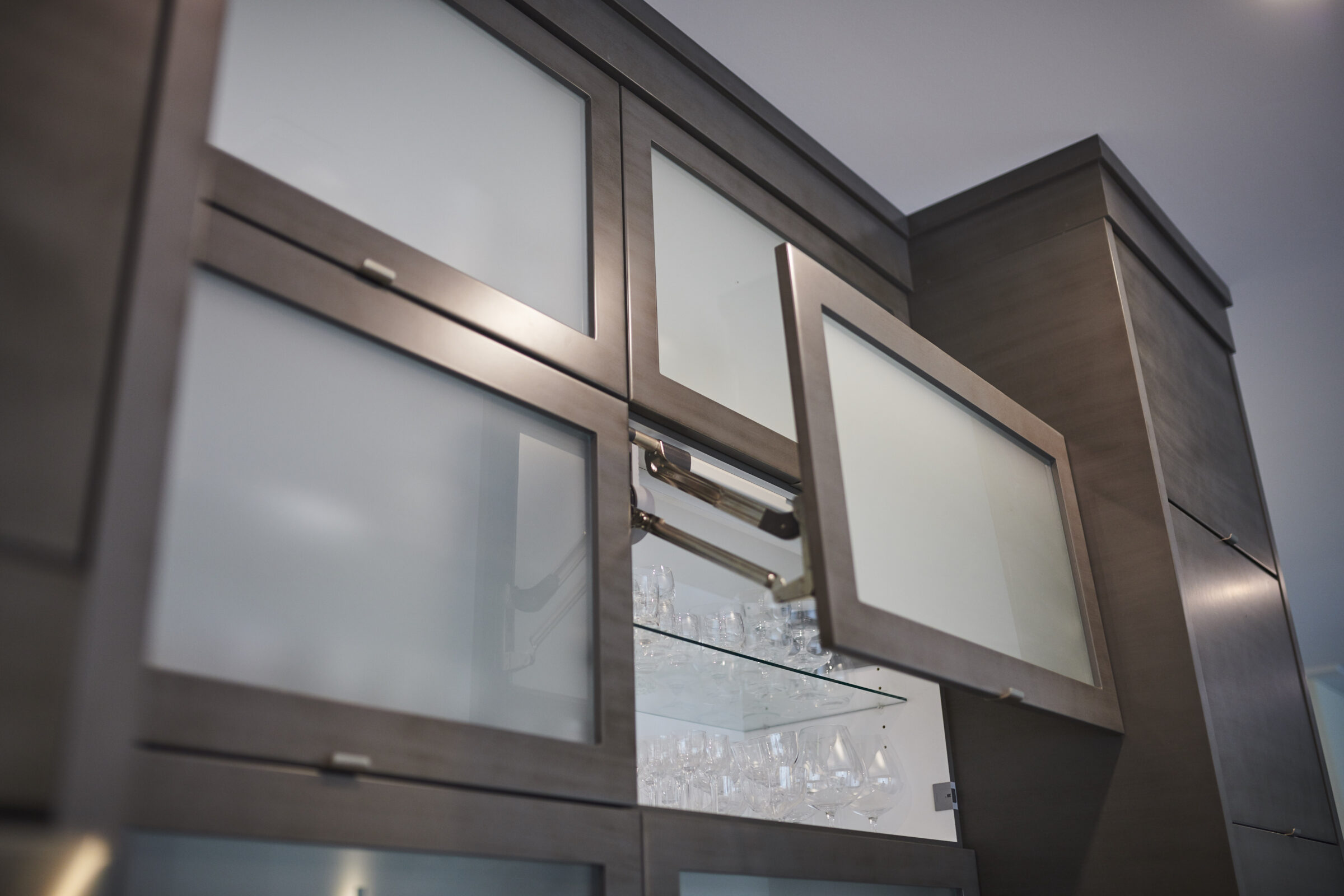 This image shows a modern kitchen with sleek, dark wood cabinets, frosted glass doors, and stainless-steel handles. An open cabinet reveals stacked glassware inside.