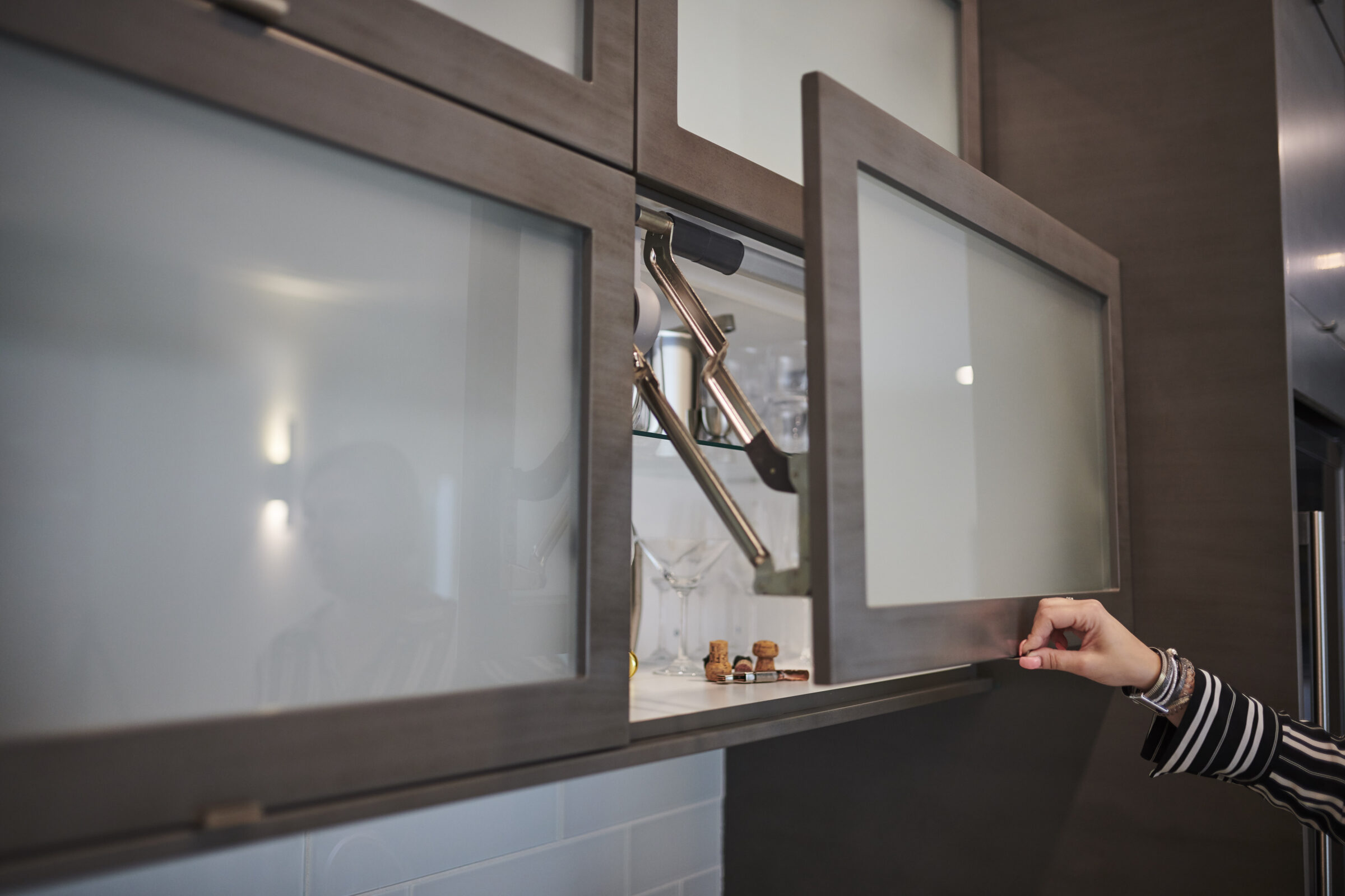 A modern kitchen cabinet with frosted glass doors is being opened by a person's hand, revealing an organized shelf with glassware inside.
