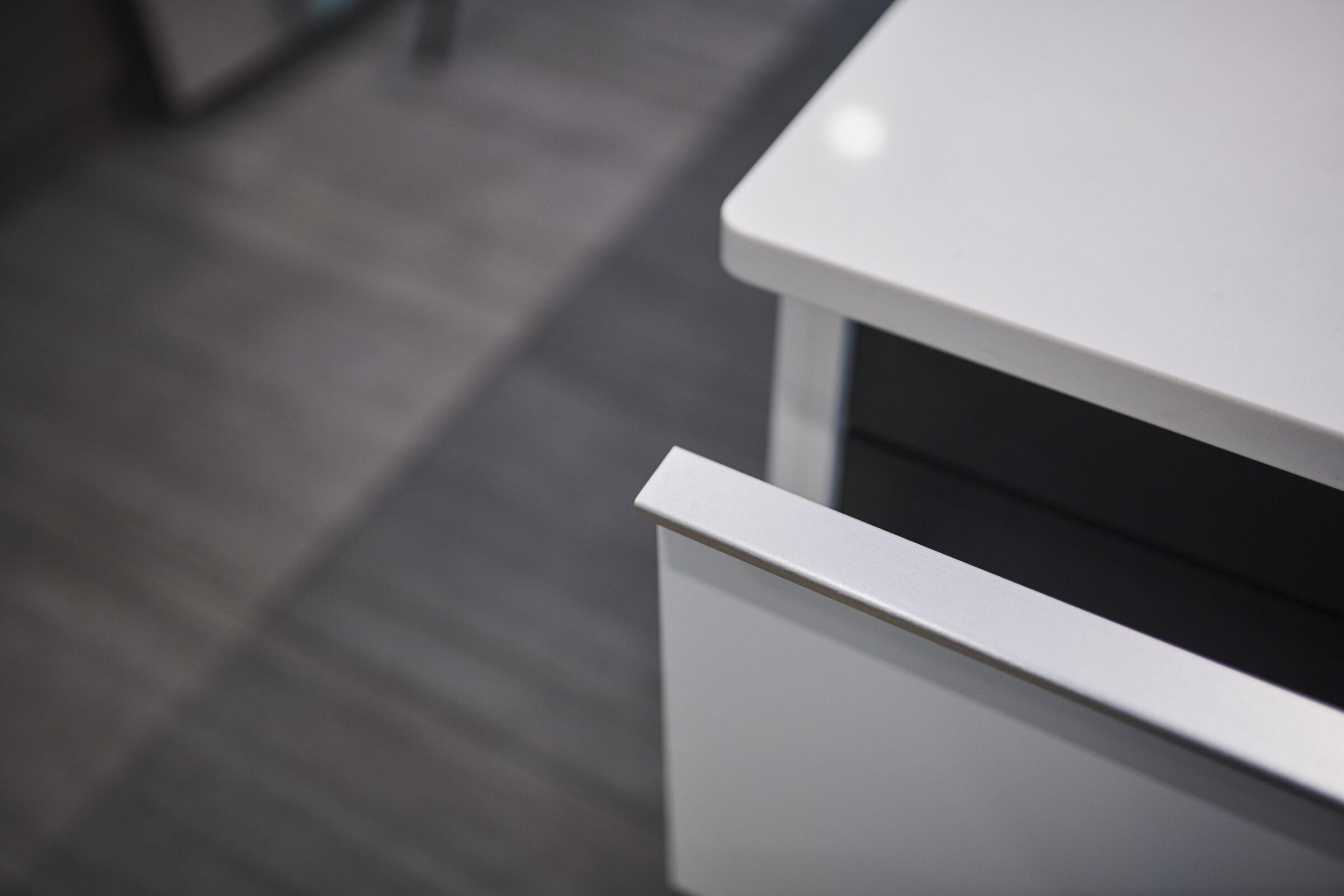 A modern, white drawer is partially open, revealing a black interior, with a blurred background showing a grey tiled floor and cabinet.