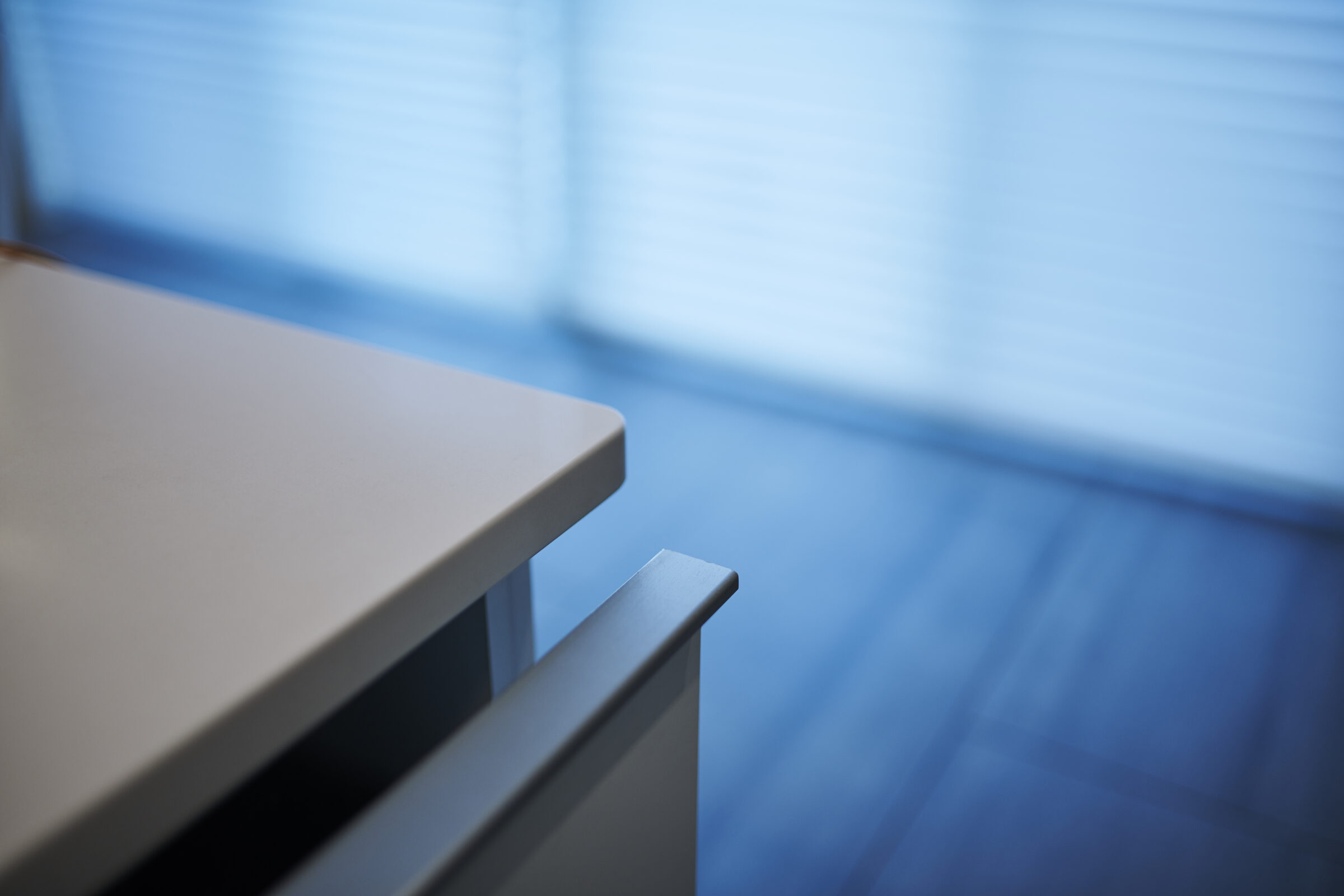 A close-up view of a modern, sleek desk corner, with a blurred background featuring blue window blinds in a softly lit room.