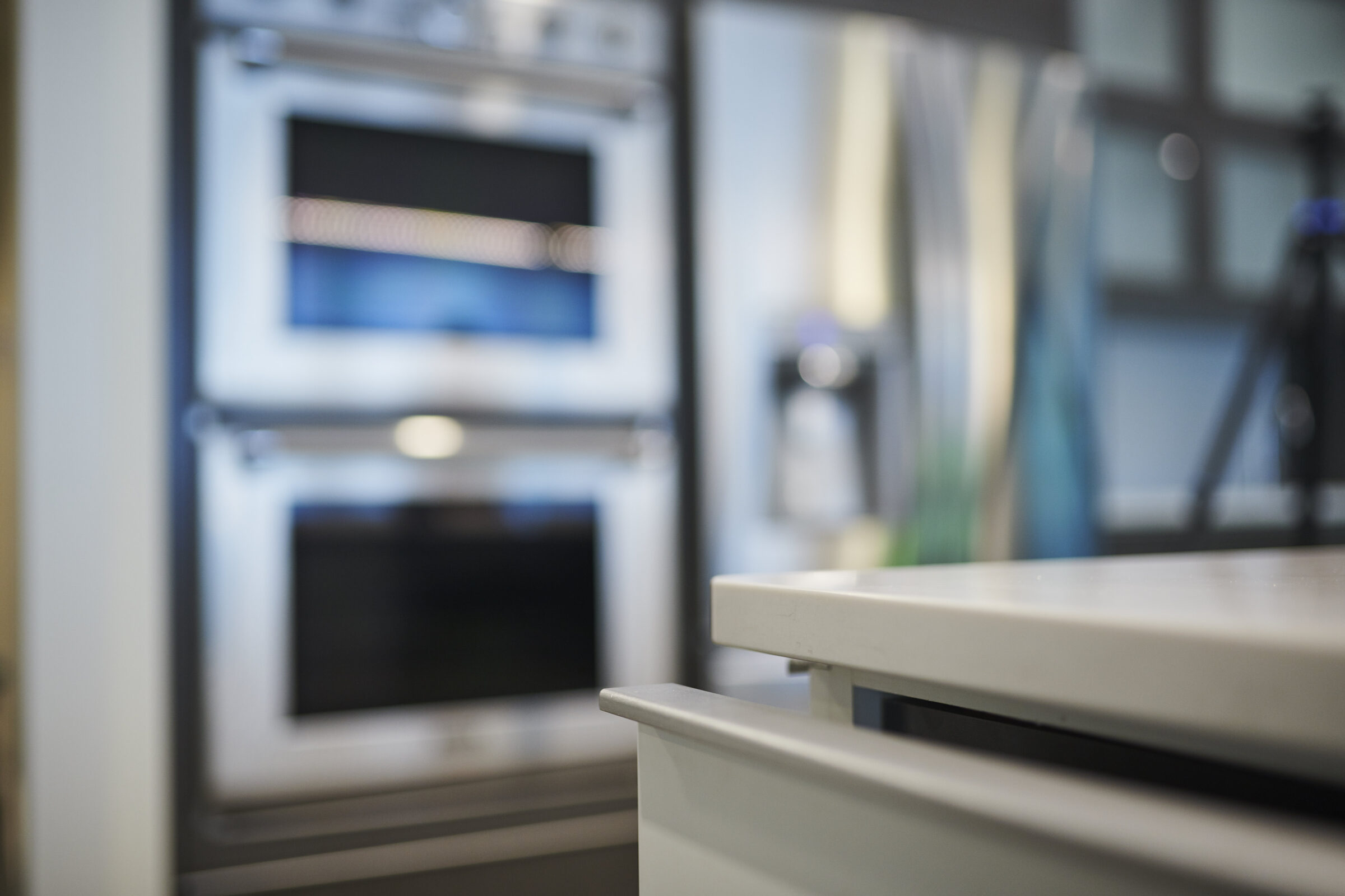 Modern kitchen interior with a focus on sleek white countertops and a blurred background showing an oven and other appliances with steel accents.