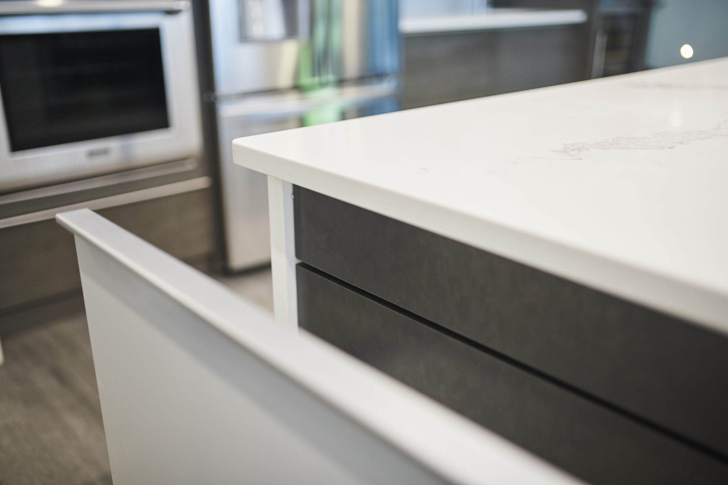 Modern kitchen close-up with a white countertop, black drawer, stainless steel microwave, and refrigerator blurred in the background. Bright, clean, minimalistic design.