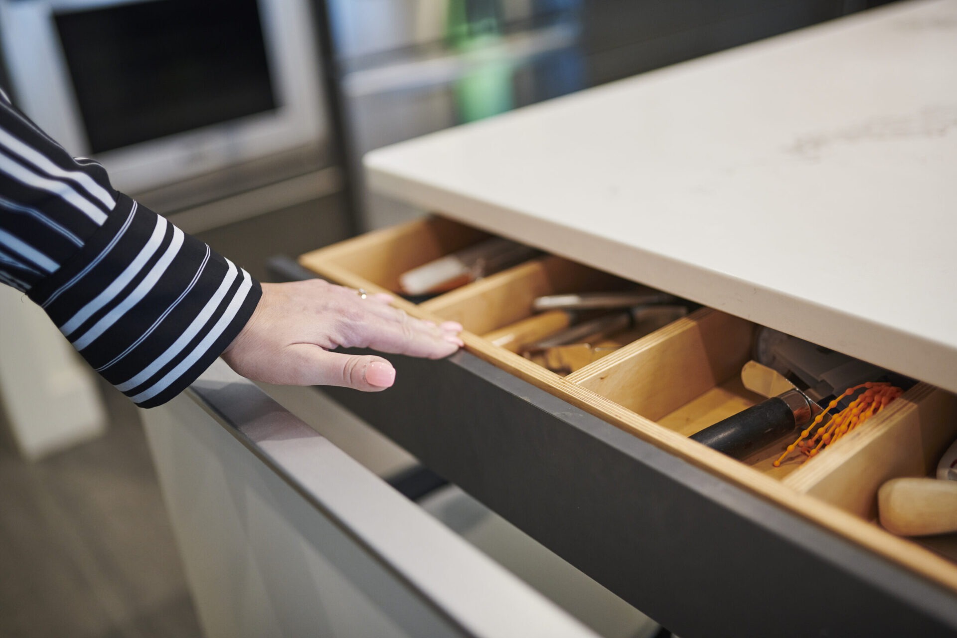 A person is opening a modern kitchen drawer revealing neatly organized utensils. The person wears a striped shirt with a ¾ sleeve.