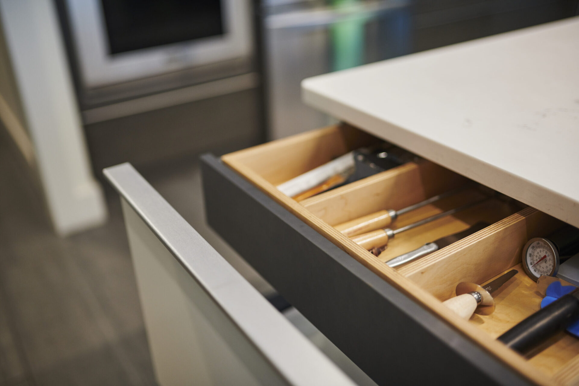 A modern kitchen drawer is partially open, revealing neatly arranged cooking utensils like knives, a timer, and other kitchen tools in an organizer.